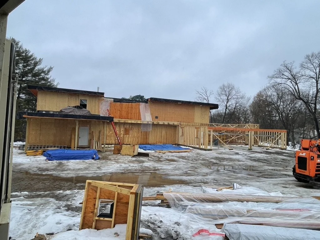 Construction site with a partially built wooden structure, some snow and ice on the ground, and construction equipment including orange lift on the right.