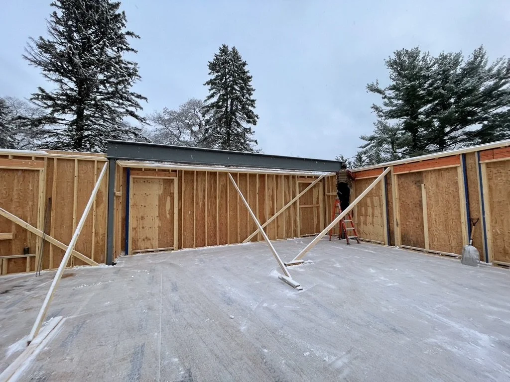 Construction site with wooden framing and support braces for a building, snowy ground, and tall trees in the background.