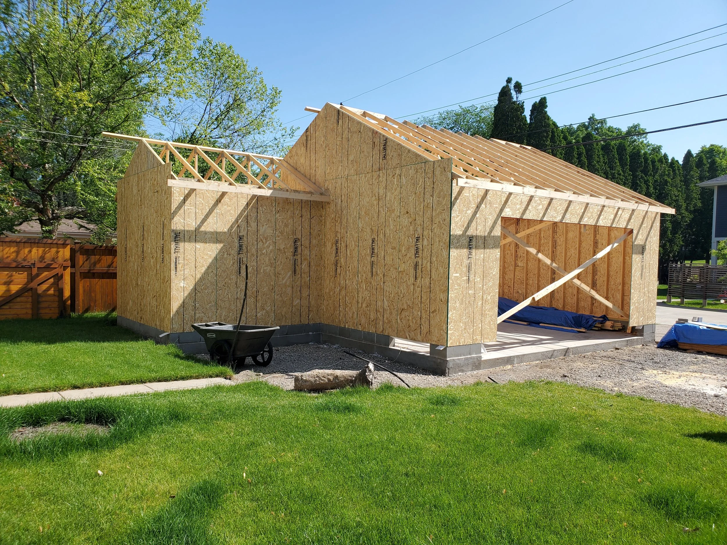 A house under construction with wooden framing and OSB sheathing, situated in a green yard with trees and a blue sky in the background.