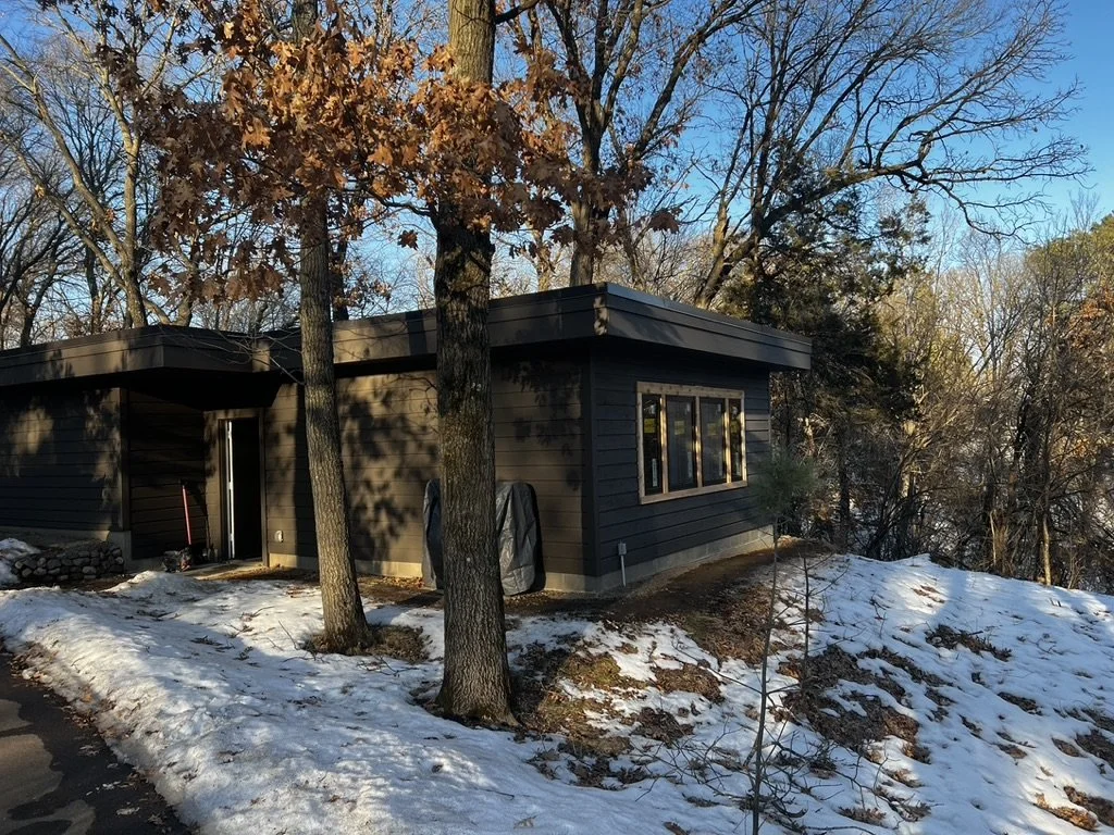 A modern black house with large windows, surrounded by trees, some with autumn leaves, and snow on the ground.