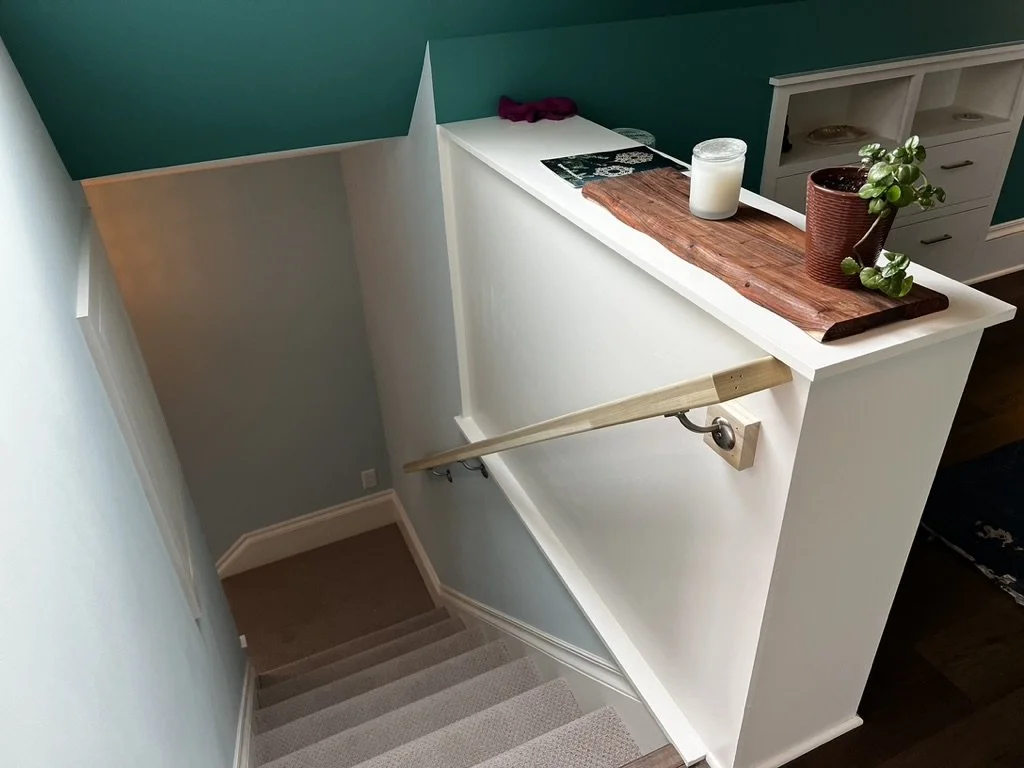 View of a staircase descending to a lower level, seen from the top, with a wooden handrail on the side and a white half wall with a ledge decorated with candles, tray, and potted plant