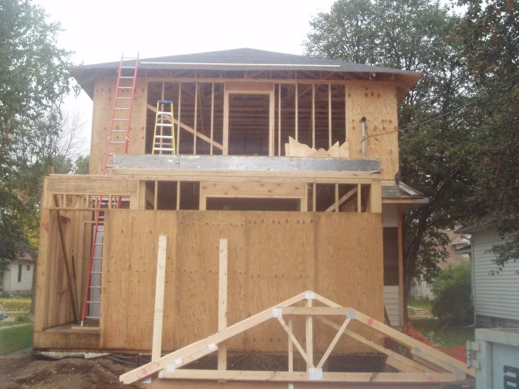 A two-story house under construction with plywood framing, ladders, and construction materials on site, surrounded by trees and nearby houses.
