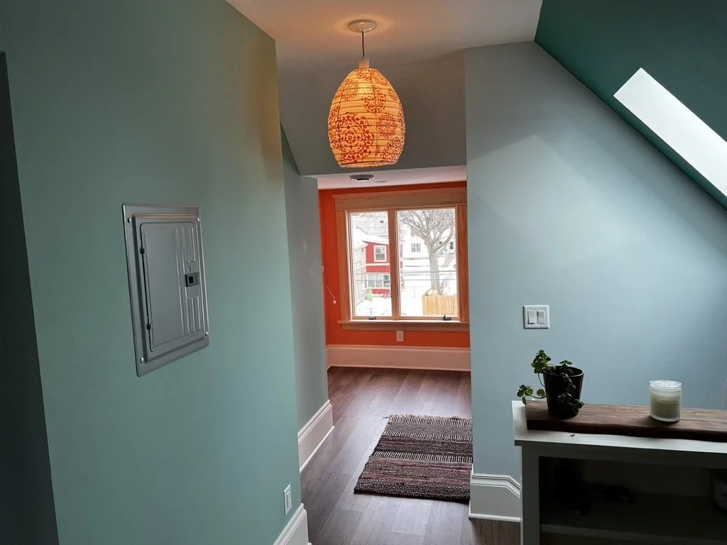 Interior of a house hallway with a view of a room featuring a large window and an orange wall, a hanging paper lantern light, a small potted plant, and a candle on a white shelf, with a striped rug on the dark wood floor.