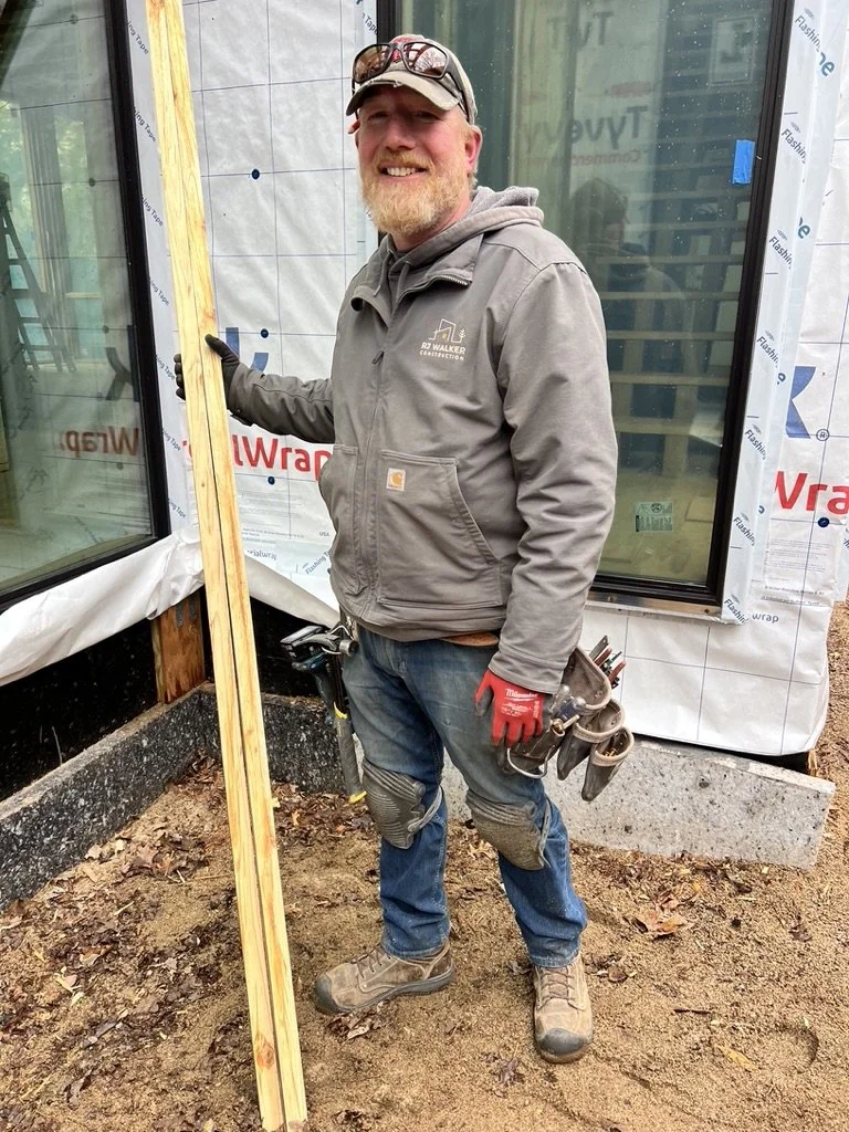 A man wearing a gray Carhartt jacket, jeans, and work boots, standing outside a building construction site. He is holding a wooden board and has a tool belt with tools, and wearing red work gloves.
