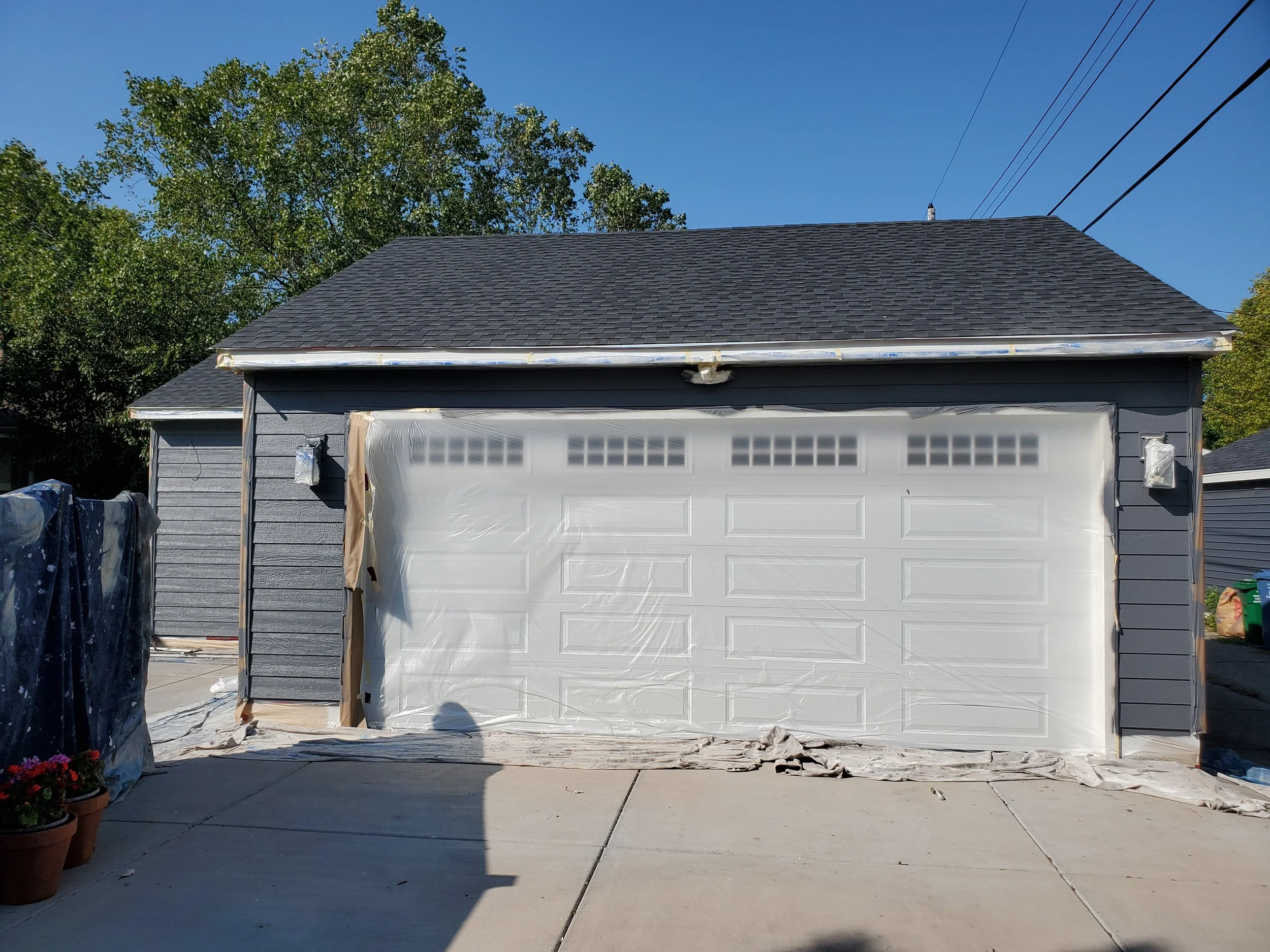 Garage door under construction with plastic covering, surrounded by protective tape, in front of a gray house with dark shingles, outdoor potted flowers, trees, and power lines in the background.