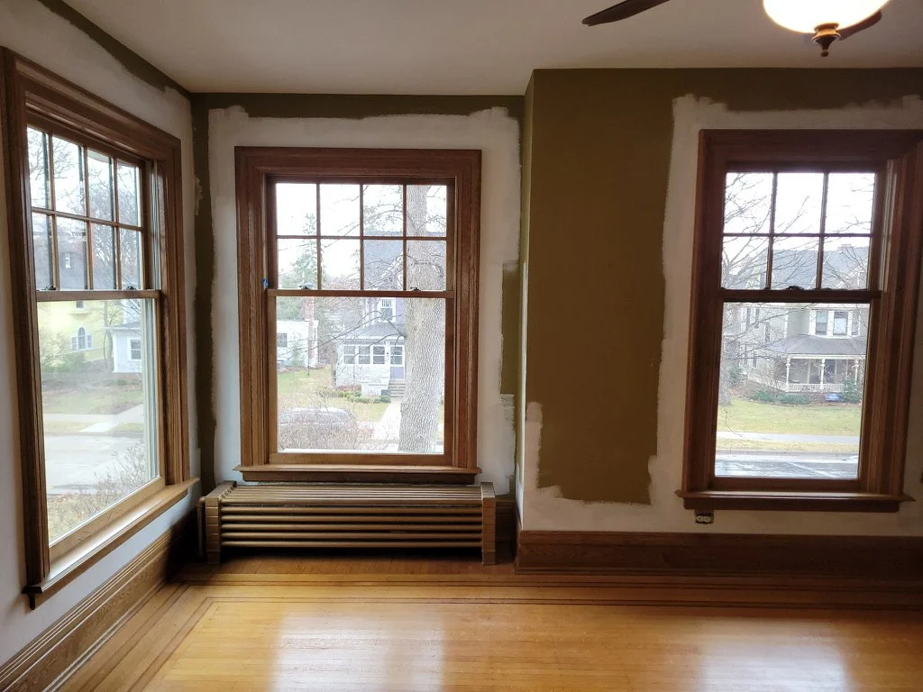 Room with three large windows, wooden frames, and a hardwood floor. Partial wall with area awaiting paint or drywall finishing. Outside view shows a residential neighborhood with houses and trees.