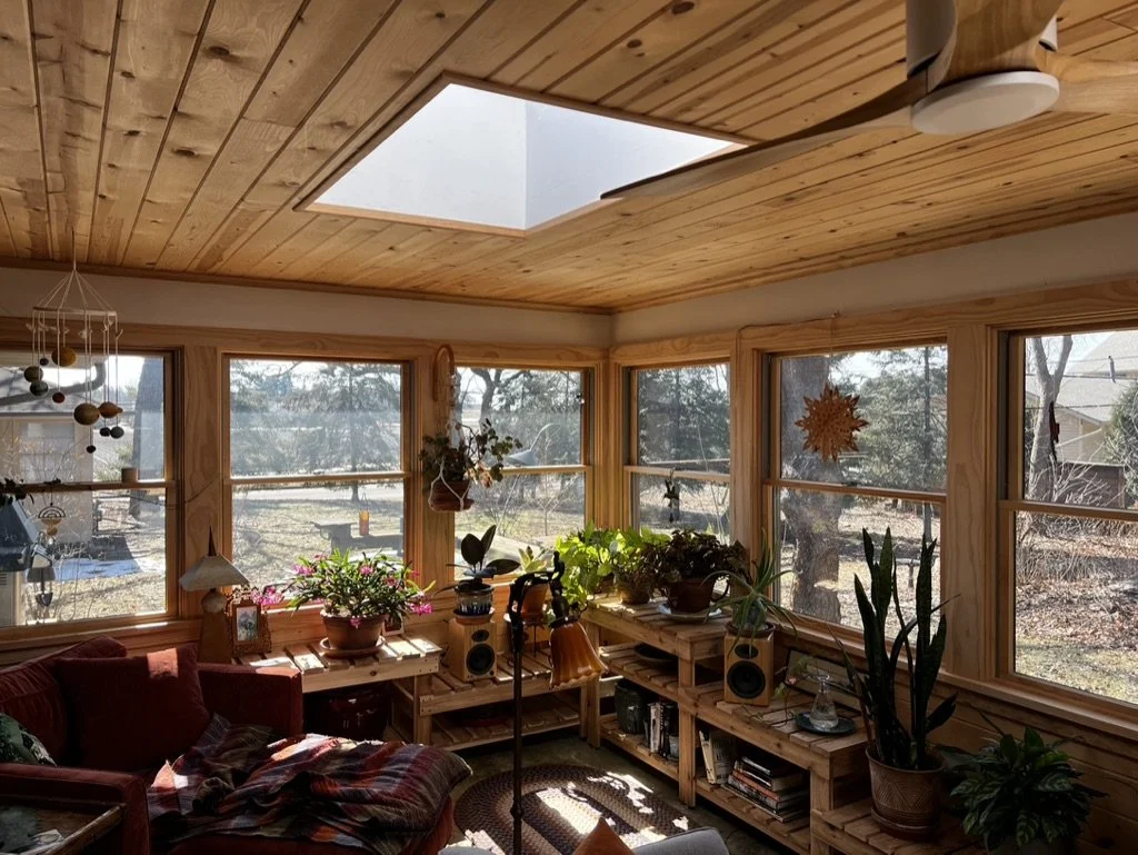Sunlit living room with wooden walls and ceiling, multiple large windows, potted plants, a red sofa, and a skylight.