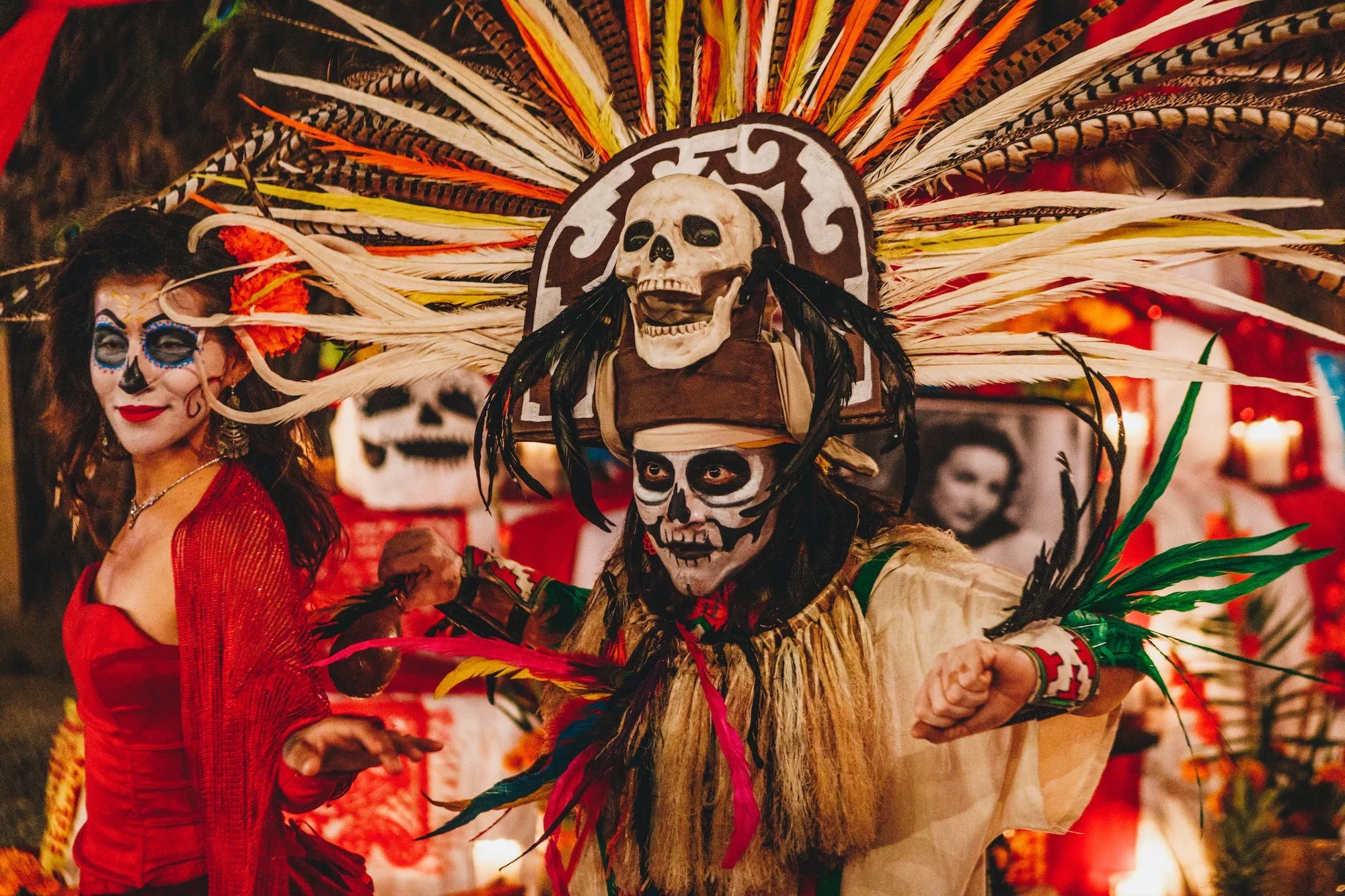 People celebrating Día de los Muertos with traditional costumes and face paint, including a woman dressed in red with sugar skull makeup and a person wearing a large headdress with skull and feather decorations.