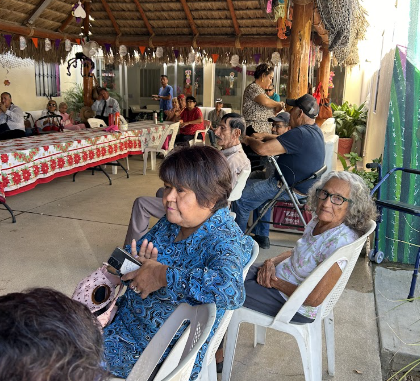 People gathered at a decorated outdoor celebration under a thatched roof, with some seated and others standing, featuring festive decorations and tables with a red and white floral tablecloth.