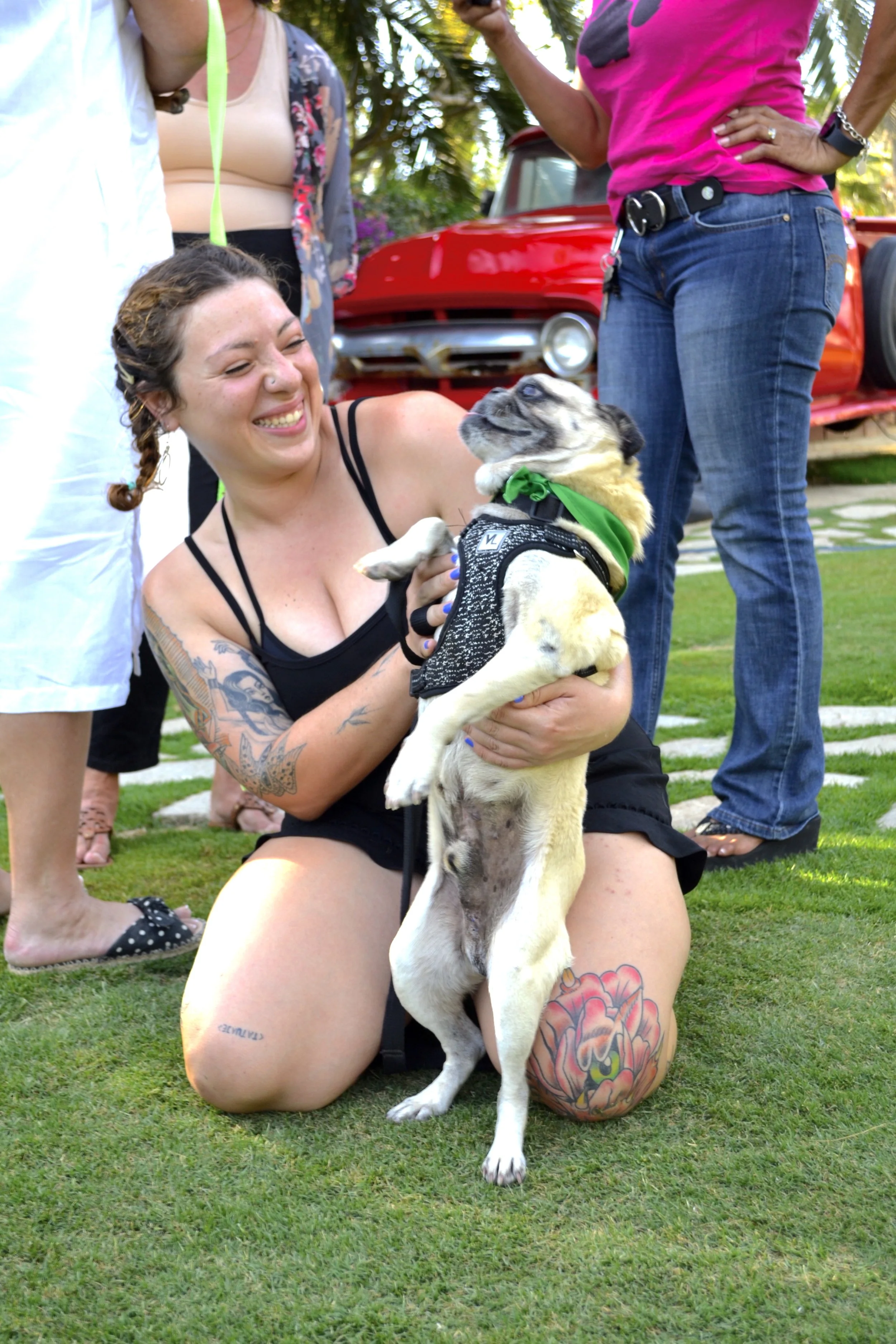 A woman with tattoos on her arms and a tattoo on her knee is kneeling on grass, holding a dog with a green bandana, surrounded by people in casual clothes in an outdoor setting.
