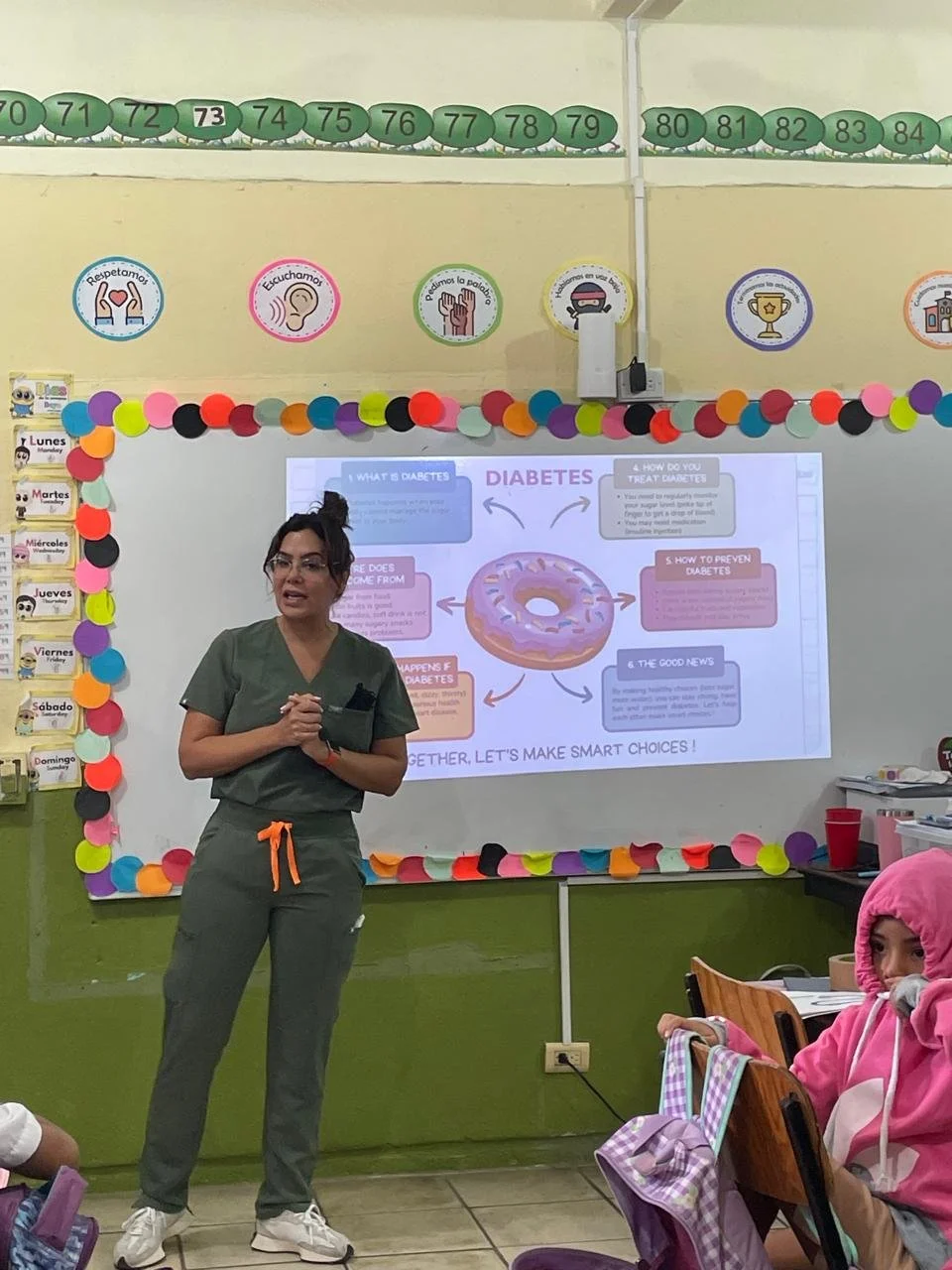 Female teacher or presenter standing in front of a classroom with a whiteboard displaying an educational slide on diabetes. Students are seated, with one girl in pink hoodie visible on the right.