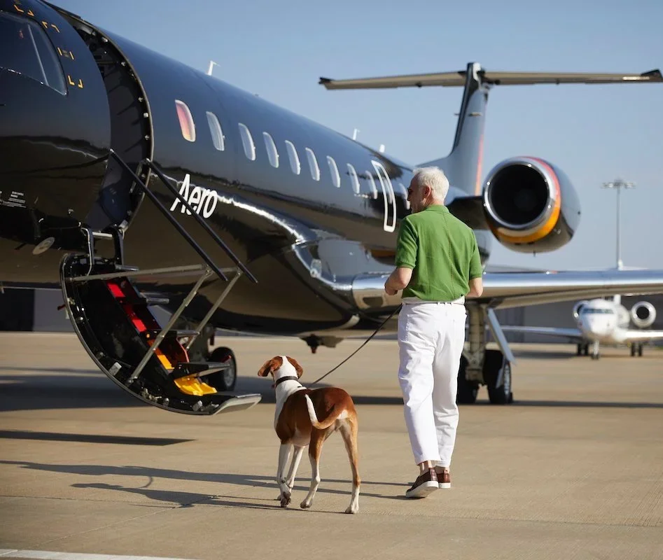 A man in a green shirt and white pants holding a dog on a leash while standing near a private jet on the tarmac.