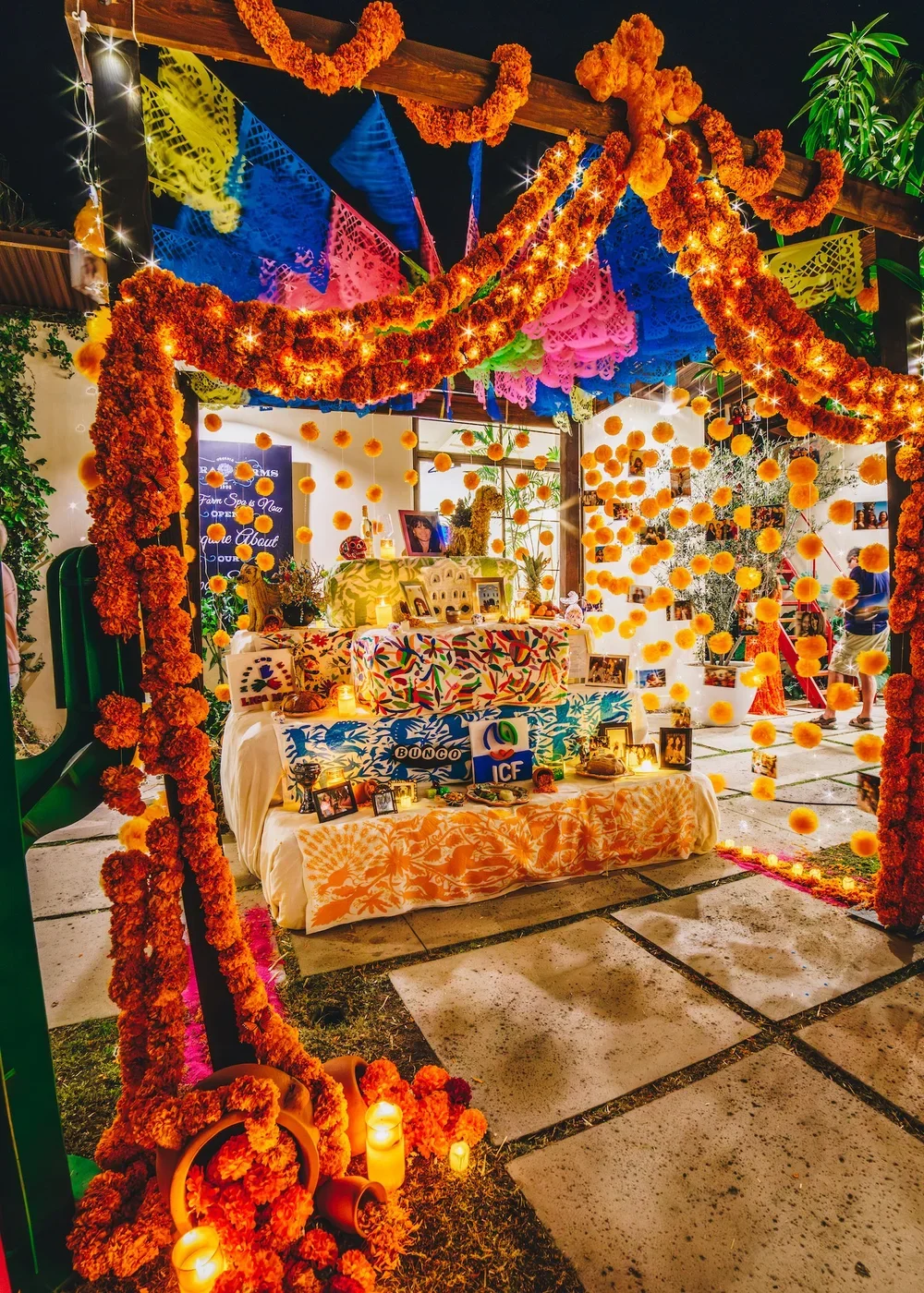 Colorful Mexican themed celebration with marigold flower decorations, papel picado banners, candles, framed photos, and a table dressed with vibrant tablecloths.
