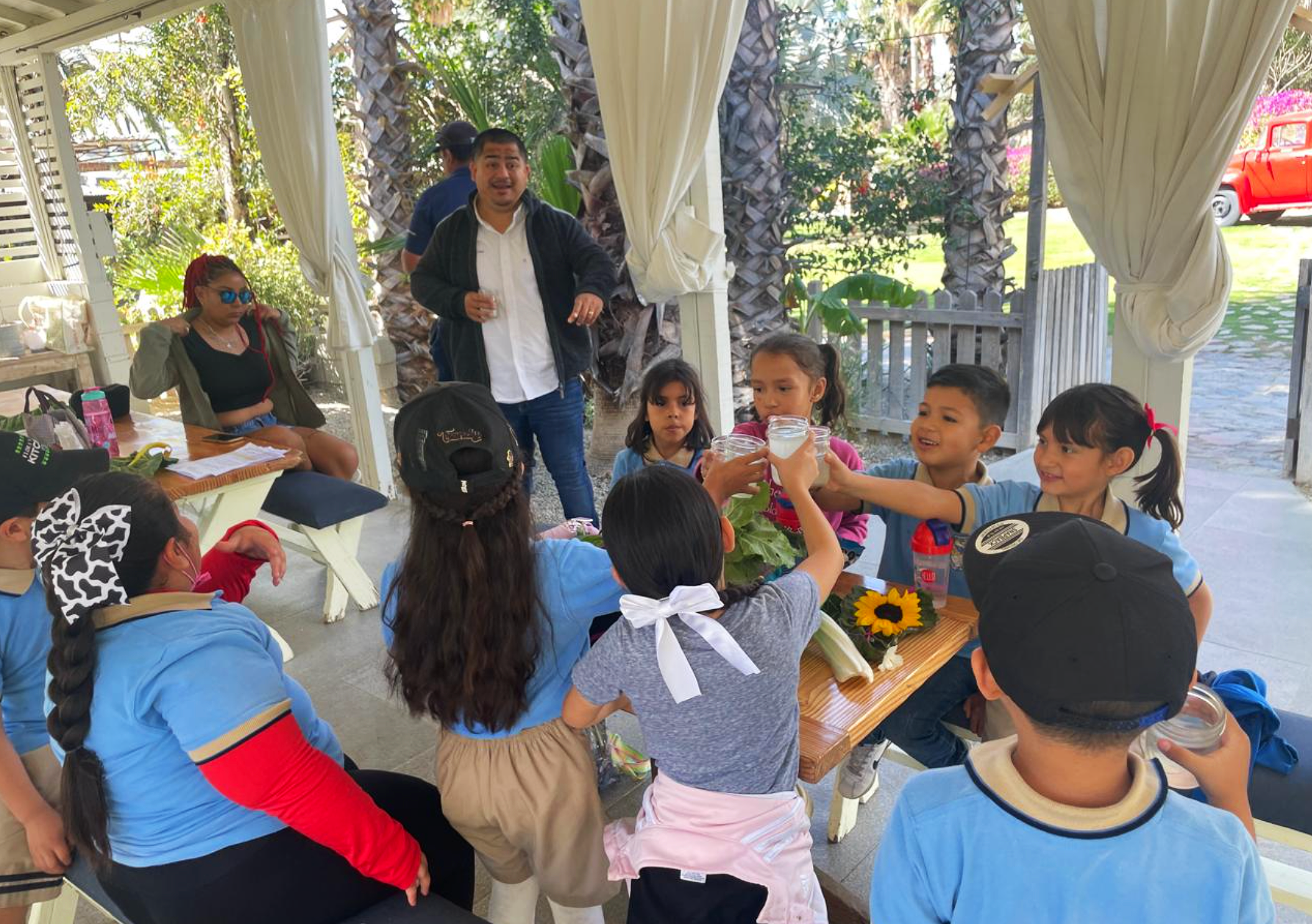 Children sitting around a table at a birthday party, with adults standing nearby, outdoors under a white canopy, with greenery and a red vehicle in the background.