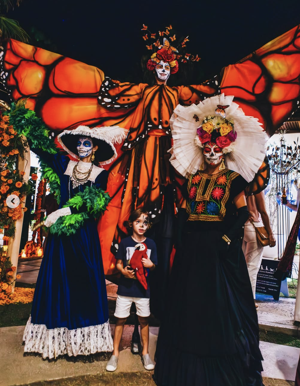 People dressed in traditional Mexican Day of the Dead costumes, including face paint and floral decorations, standing with a young boy.