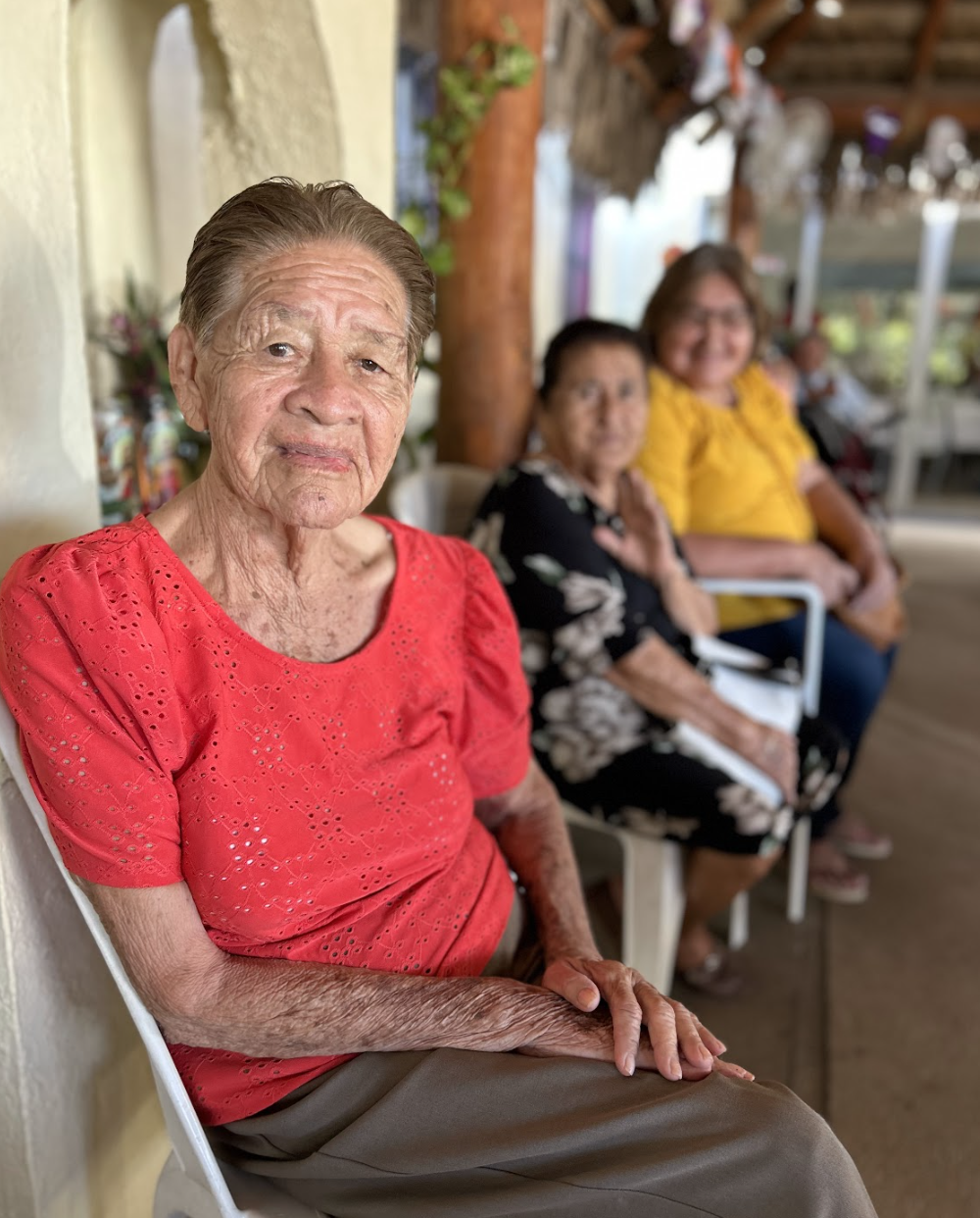 An elderly woman sitting on a white chair wearing a red blouse with three other women sitting behind her, all in an outdoor or semi-outdoor setting.