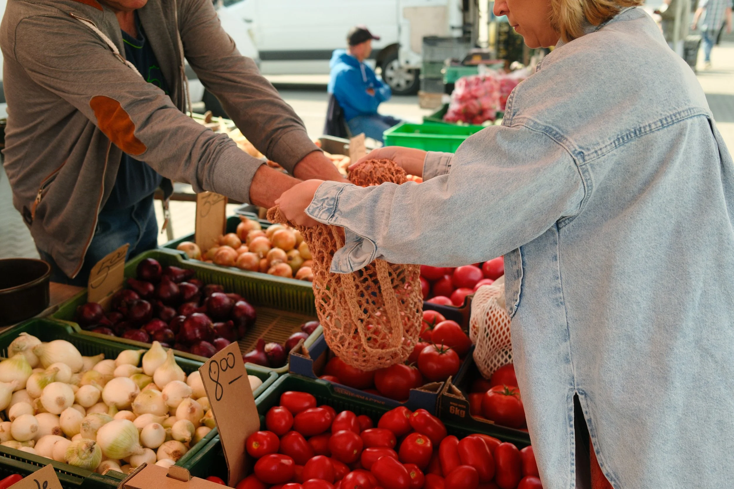 Two people exchanging money at an outdoor farmers market with baskets of onions, tomatoes, and other produce.