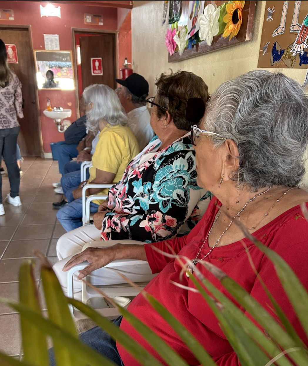 A waiting room at a restaurant with elderly women seated in chairs, one wearing a red top and glasses, another in a floral blouse, and others in casual attire. Some are engaged in conversation or looking around. The room has colorful wall art and a small sink in the corner, with a woman standing near the door.