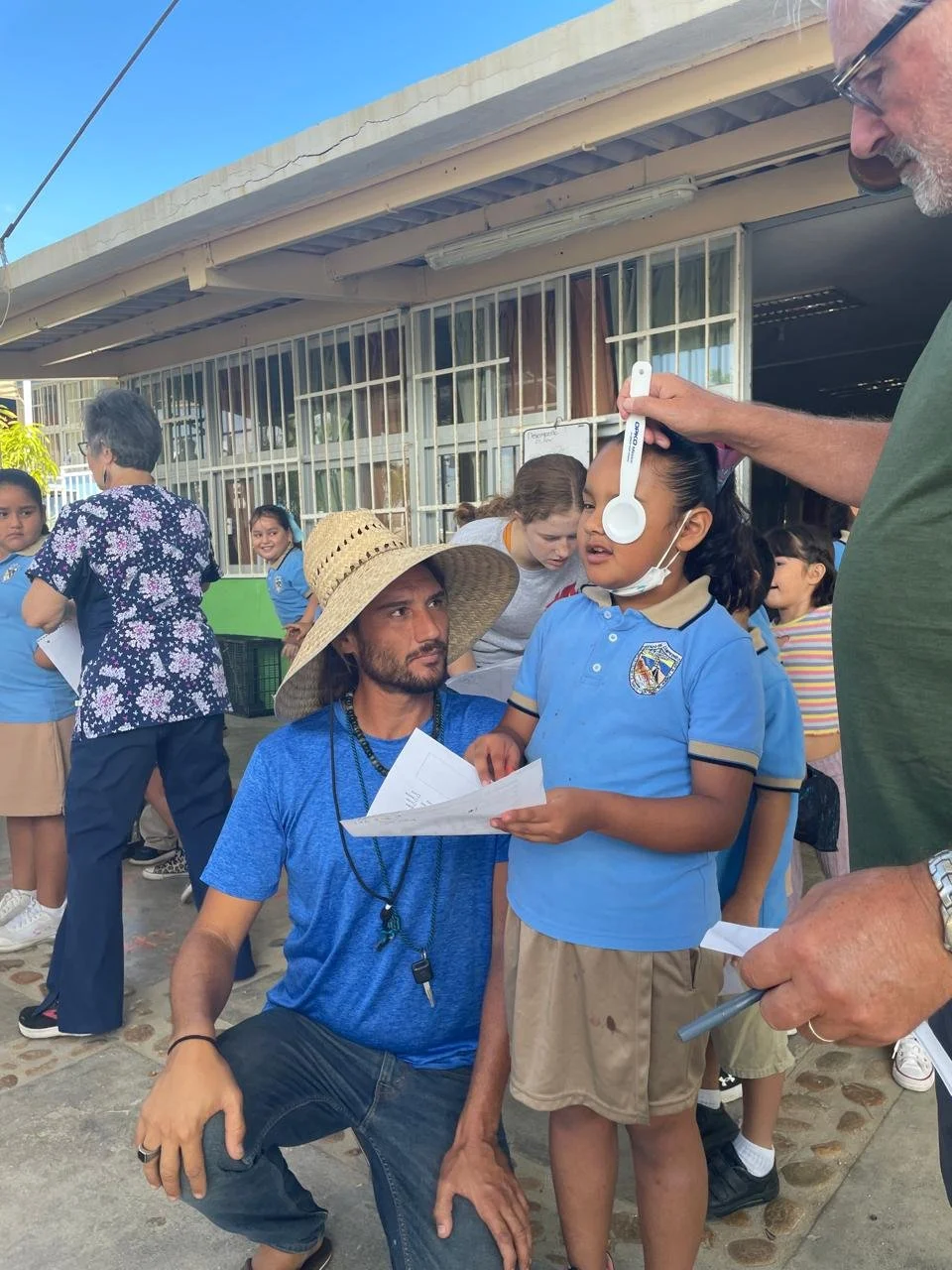 A young girl in a blue school uniform and glasses having her ears examined with an otoscope by a man, with a group of children and adults in the background outside a school building.