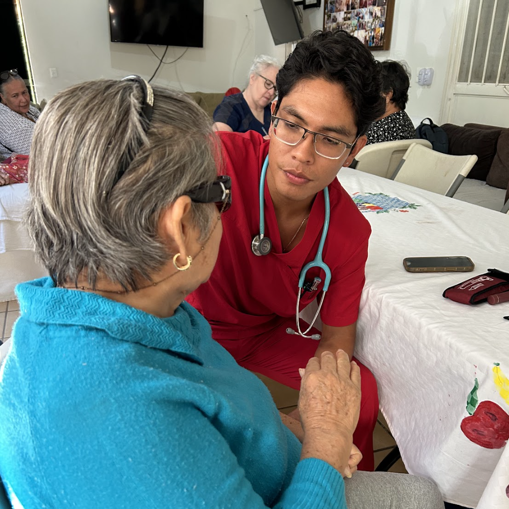 A young healthcare professional in red scrubs and glasses listens attentively to an elderly woman with gray hair and glasses, seated at a table with a white tablecloth. They are holding hands and engaging in a caring conversation in a communal setting.