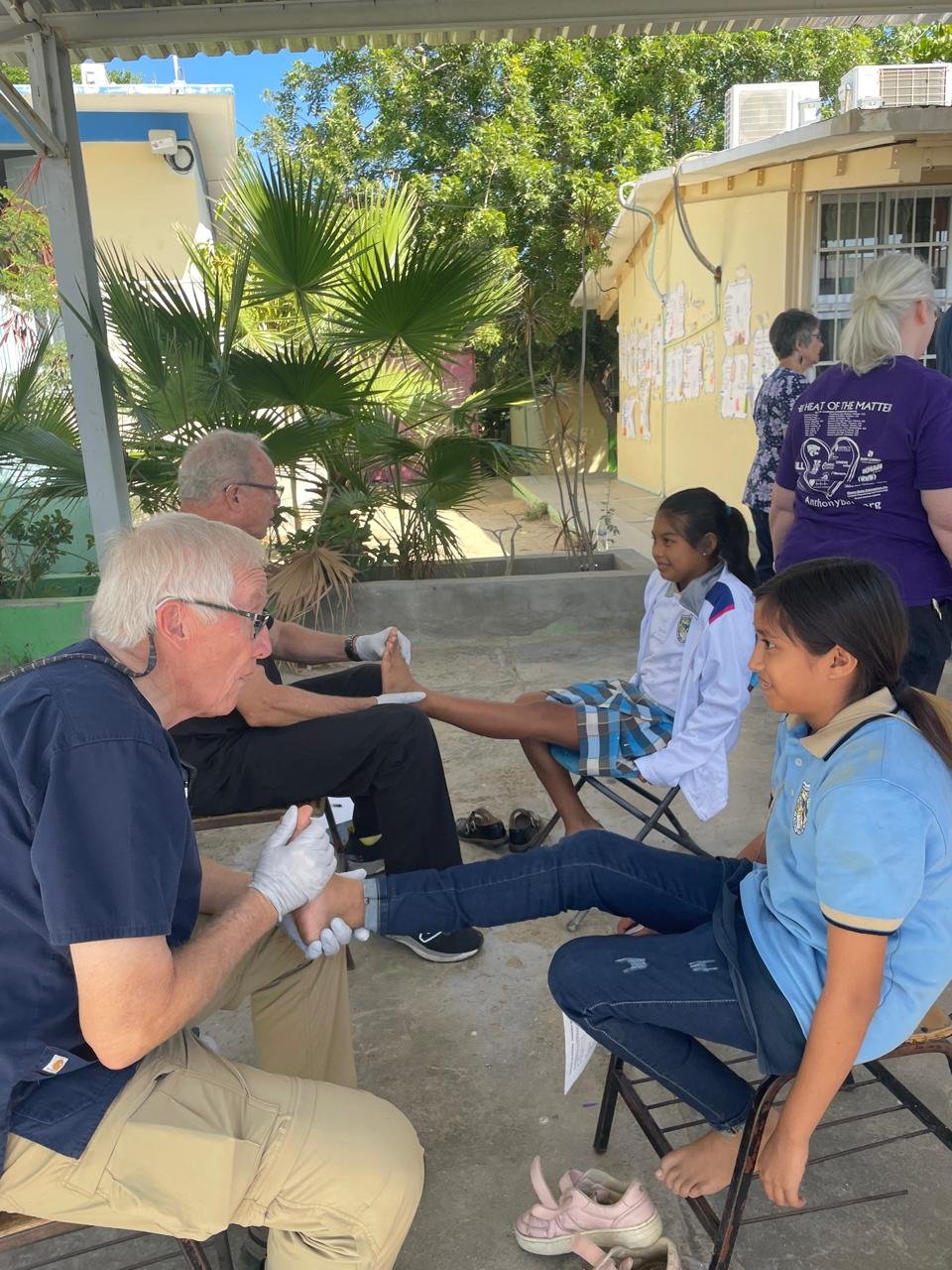 Two elderly men and two young girls at a school outdoor setting, with the girls receiving foot care and the men providing assistance. There are plants and a yellow building in the background.