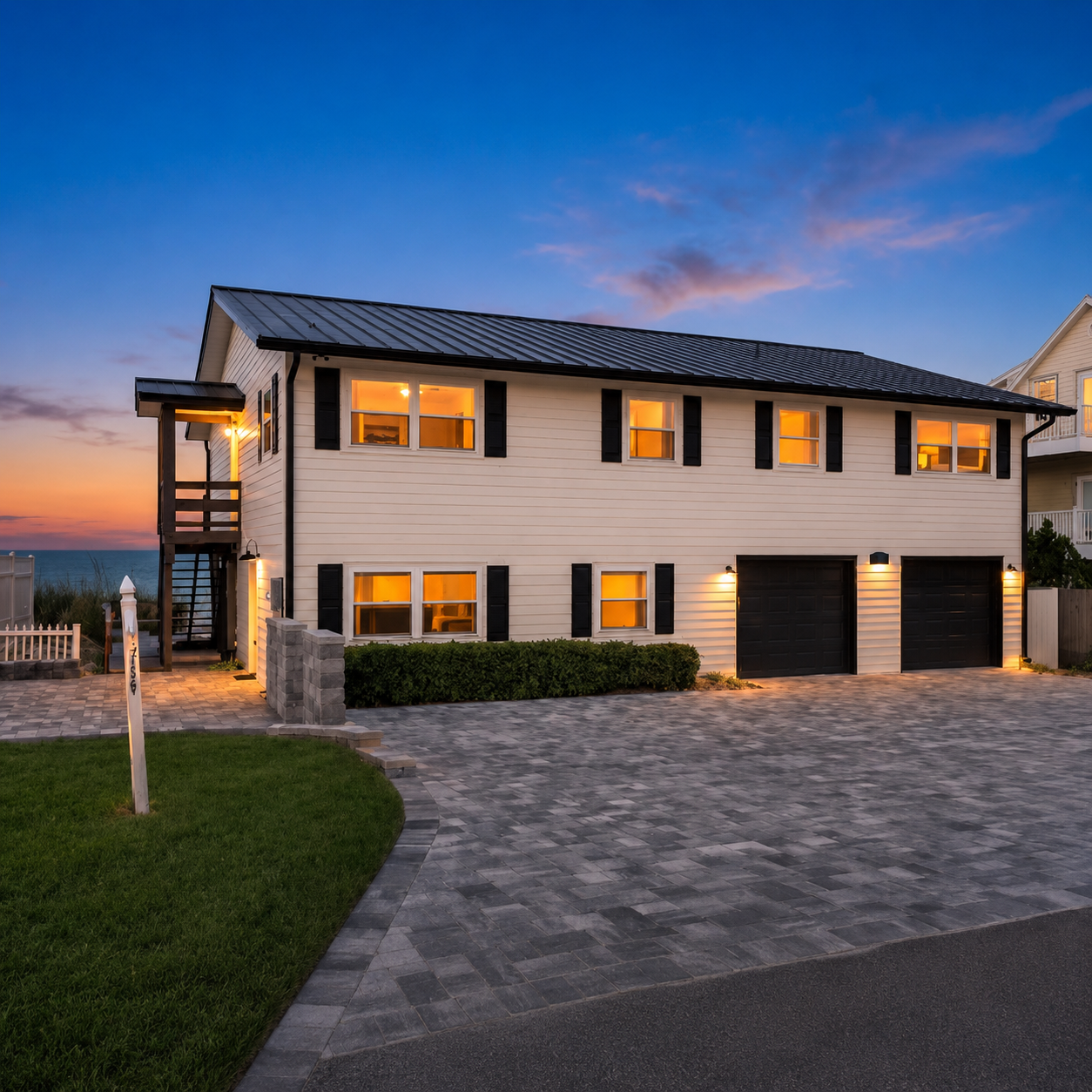 Modern two-story house with black window shutters, attached garage, and exterior lighting at sunset, overlooking a body of water.