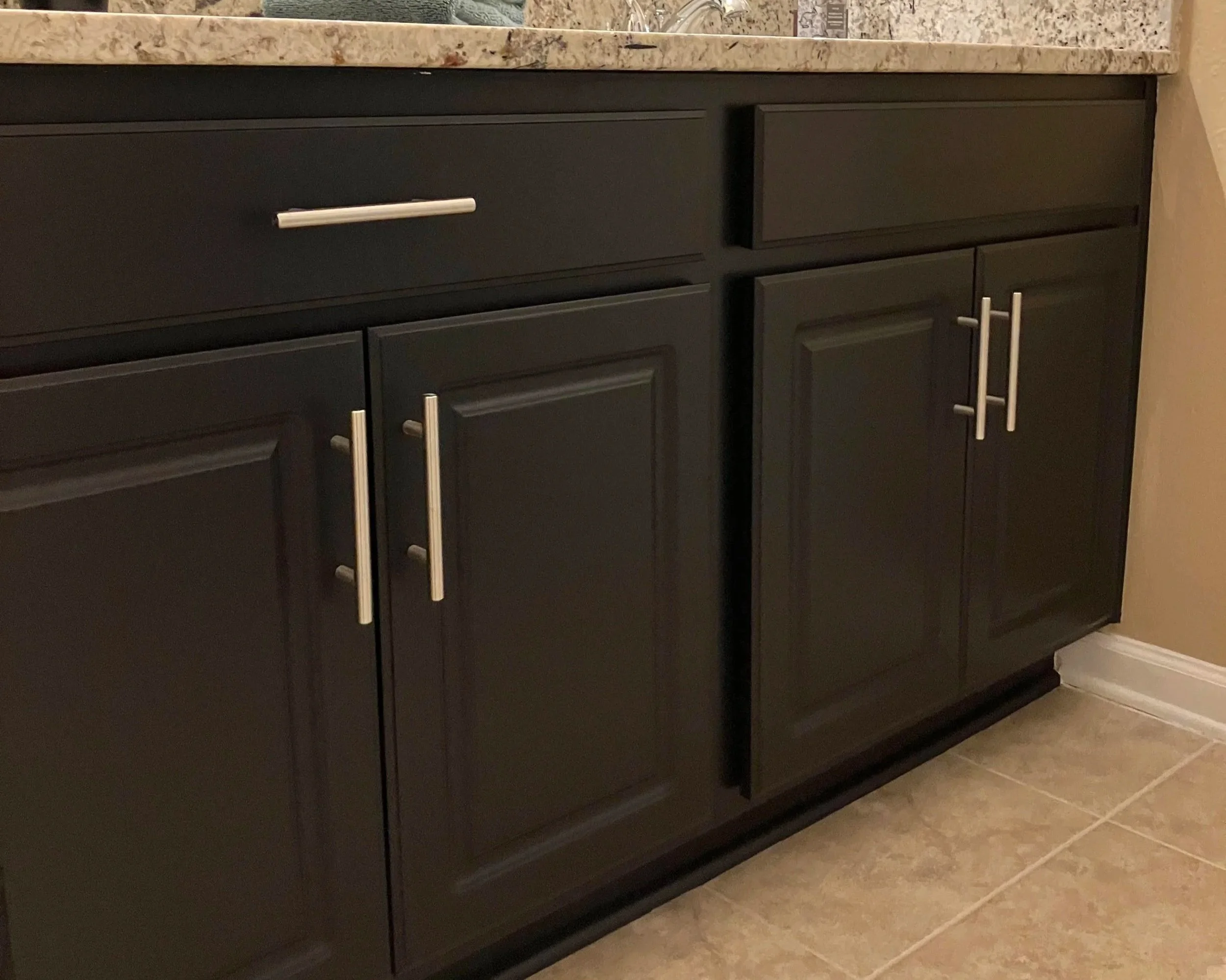 A black kitchen cabinet with silver handles and a granite countertop, on a beige tiled floor.