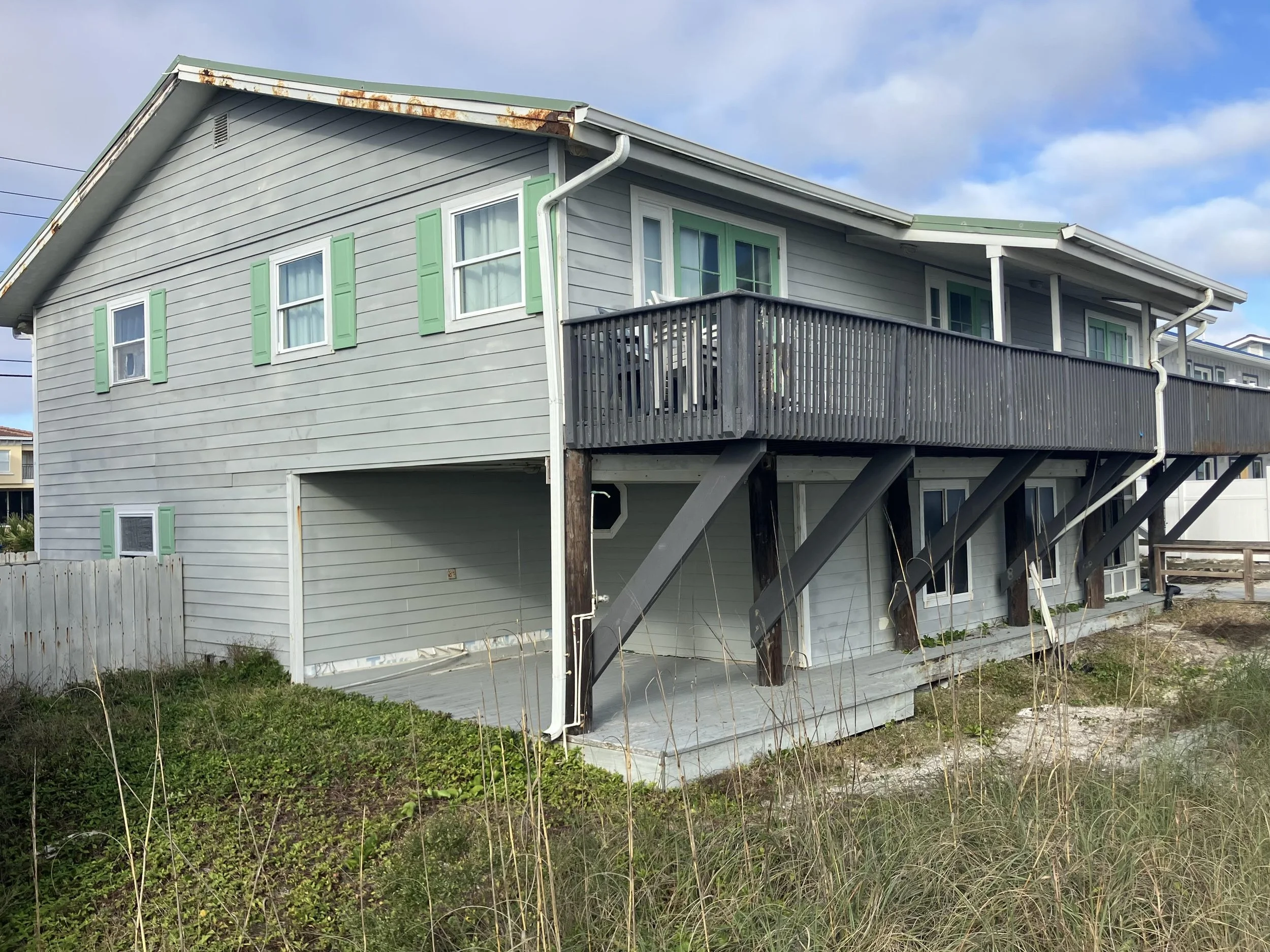 A two-story house with light gray siding and green shutters, featuring a deck on the upper level and a fenced yard below. The house shows signs of rust on the roof edge, and the yard has some overgrown grass and weeds.