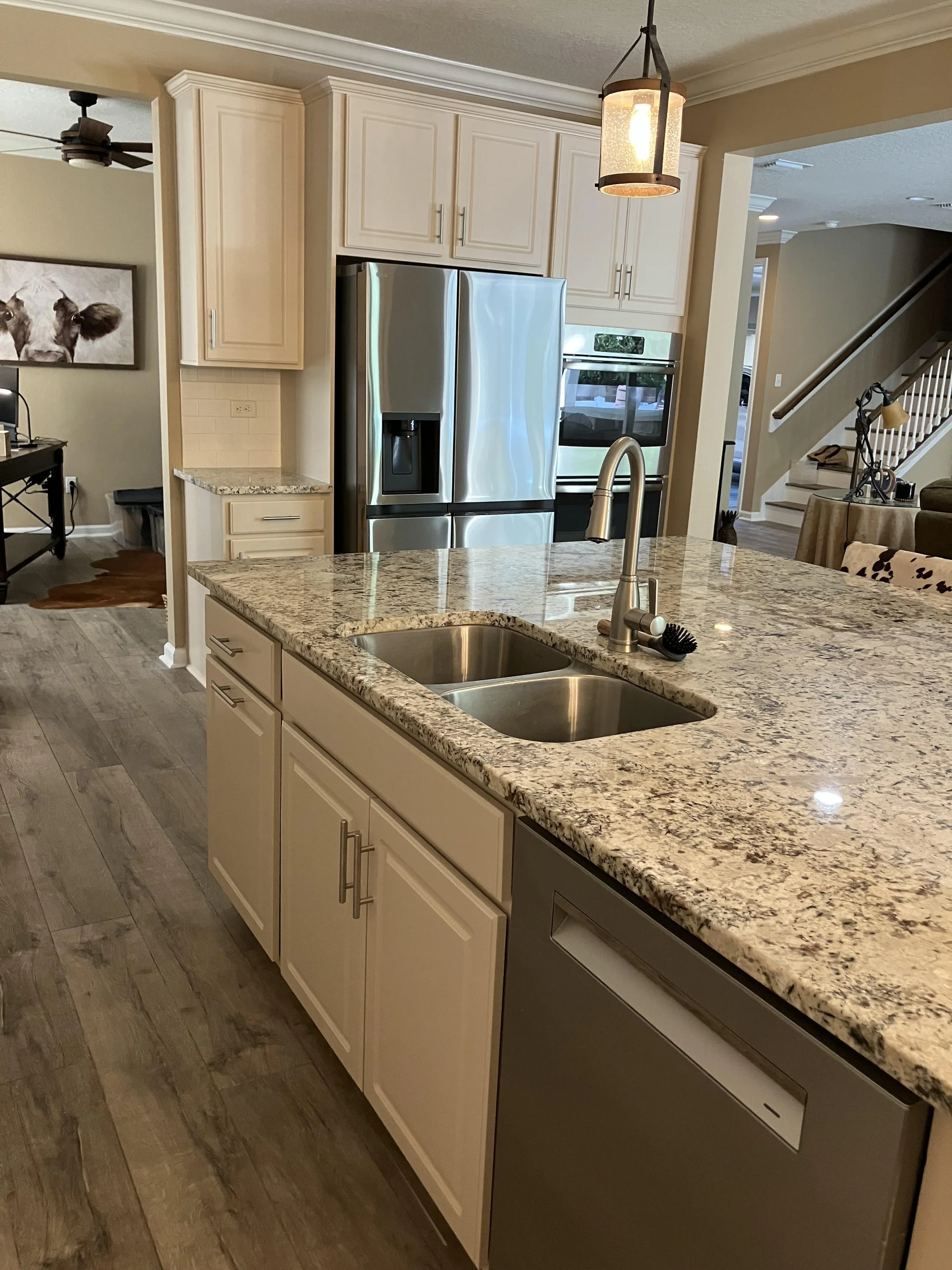 A modern kitchen with a granite countertop island, stainless steel refrigerator, and built-in oven. There is a pendant light hanging above the island and a sink with a faucet. The kitchen has light-colored cabinets and wood flooring.
