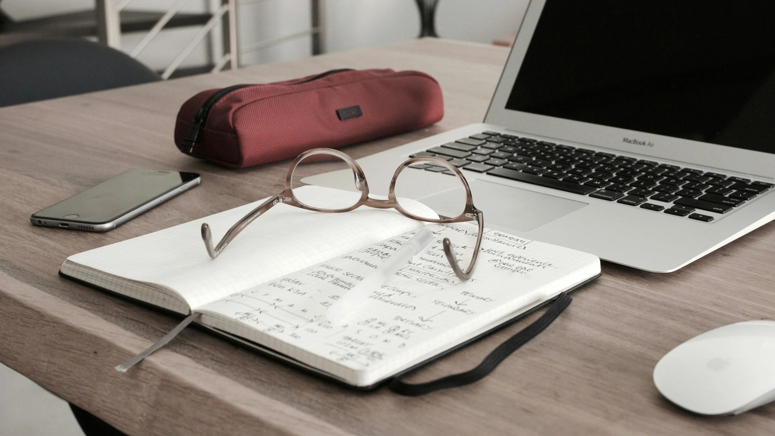 A desk with a pair of glasses, a notebook filled with handwritten notes and a pen, a closed laptop, a smartphone, a red pencil case, and a white computer mouse.