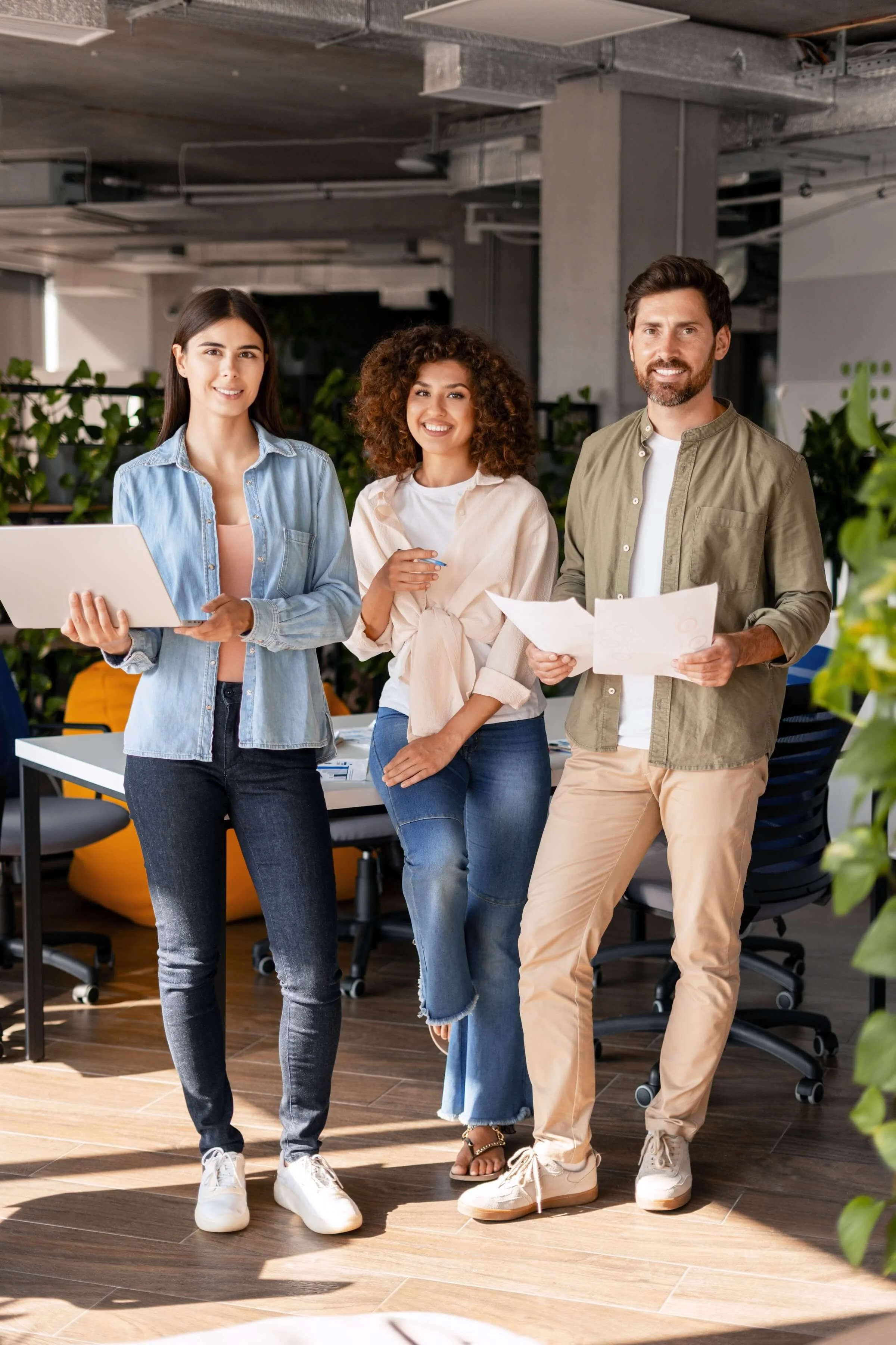 Three young professionals standing in an office, smiling, with a woman holding a tablet and the man holding documents.