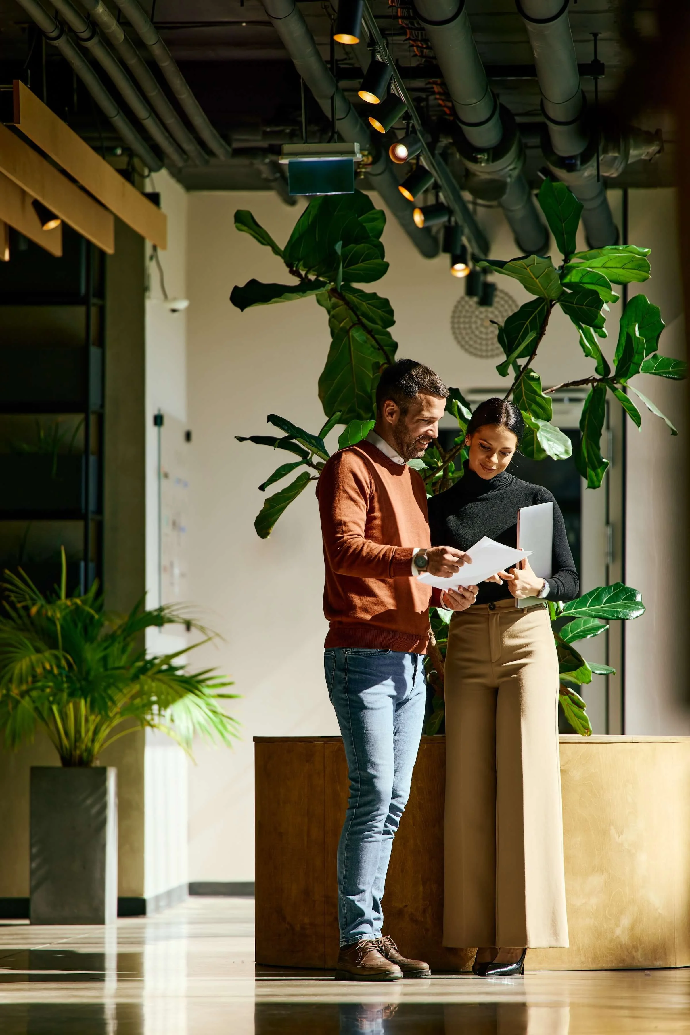 A man and woman in business casual attire are standing together indoors, looking at and discussing a piece of paper, with a laptop in the woman's hands. They are standing next to a large green leafy plant, and the background features a modern space with overhead pipes and lighting.