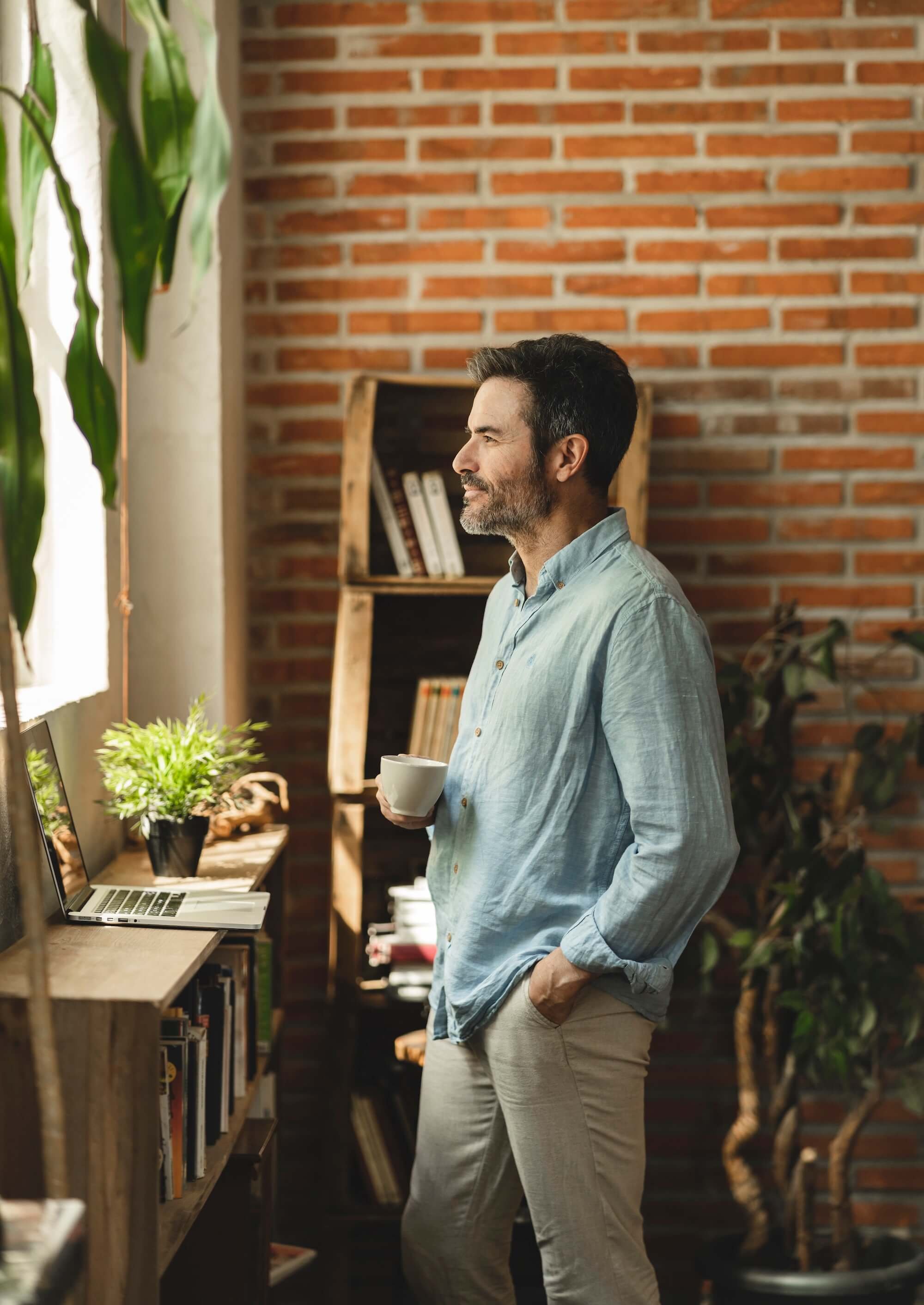 A man with dark hair and a beard, dressed in a light blue shirt and beige pants, stands indoors holding a cup of coffee, gazing out the window with a thoughtful expression. The room features a brick wall, bookshelf, potted plant, and a laptop on the wooden shelf.