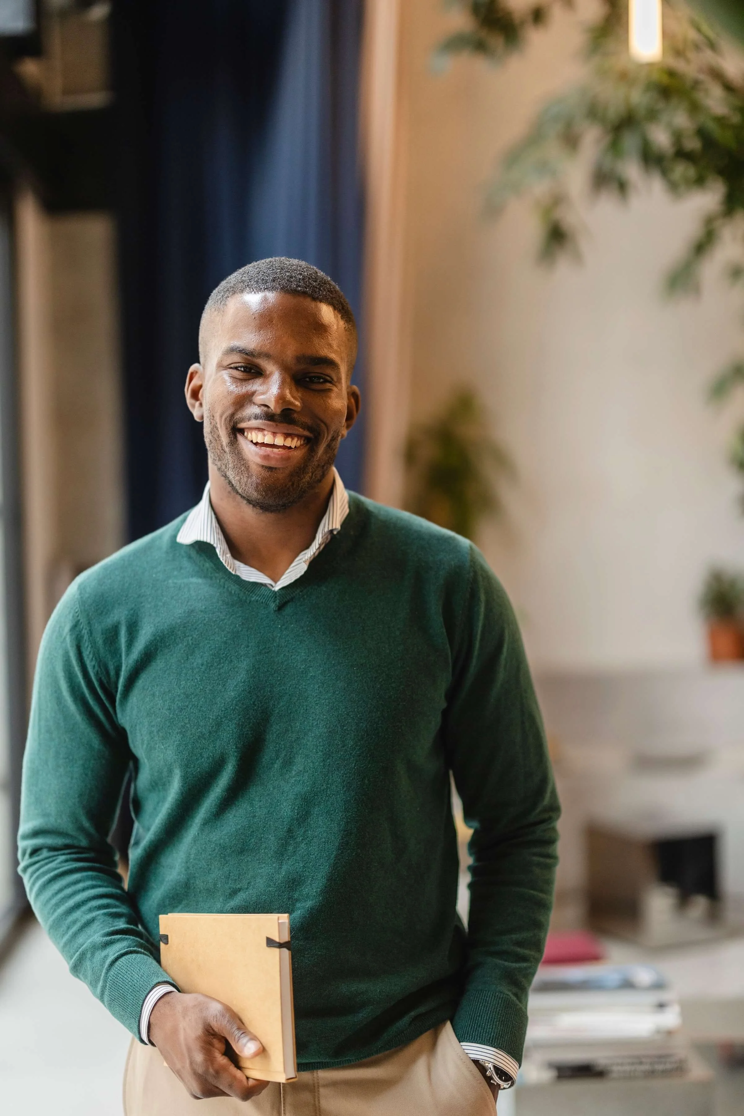 A smiling man in a green sweater and collared shirt stands indoors, holding a notebook.