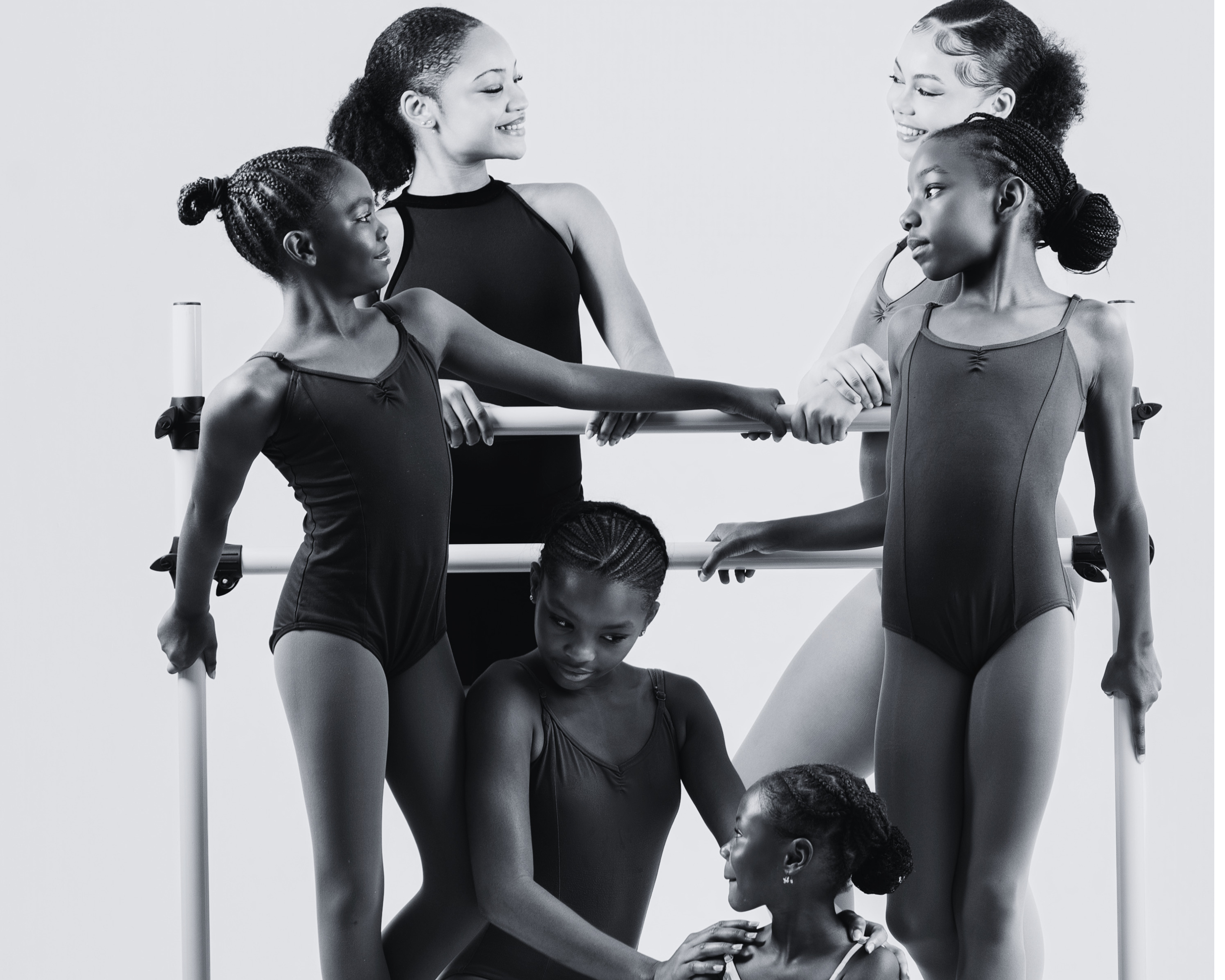Five young female gymnasts with two adult women in a black and white photo. The girls are wearing leotards, and the women are in athletic wear. They are smiling and holding a horizontal bar.