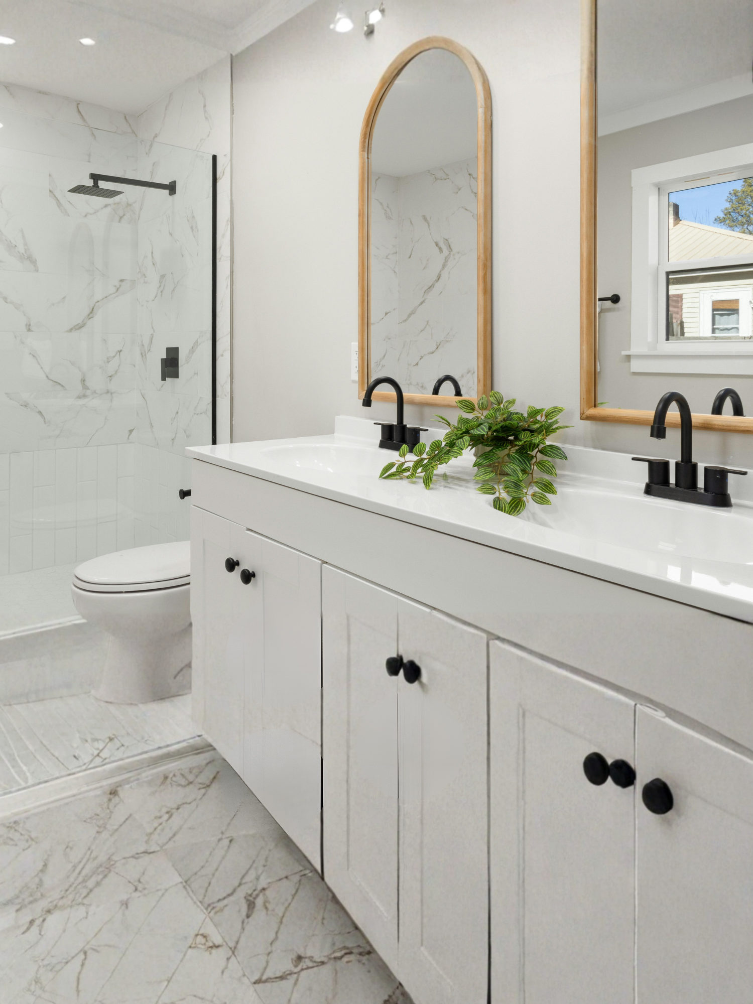 Modern bathroom with a white vanity, black fixtures, and two arched mirrors. A wood-framed mirror is above the sink with a plant on the countertop. The shower area features white marble tiles with gray veining and a black rainfall showerhead. A small window shows part of a house and trees outside.
