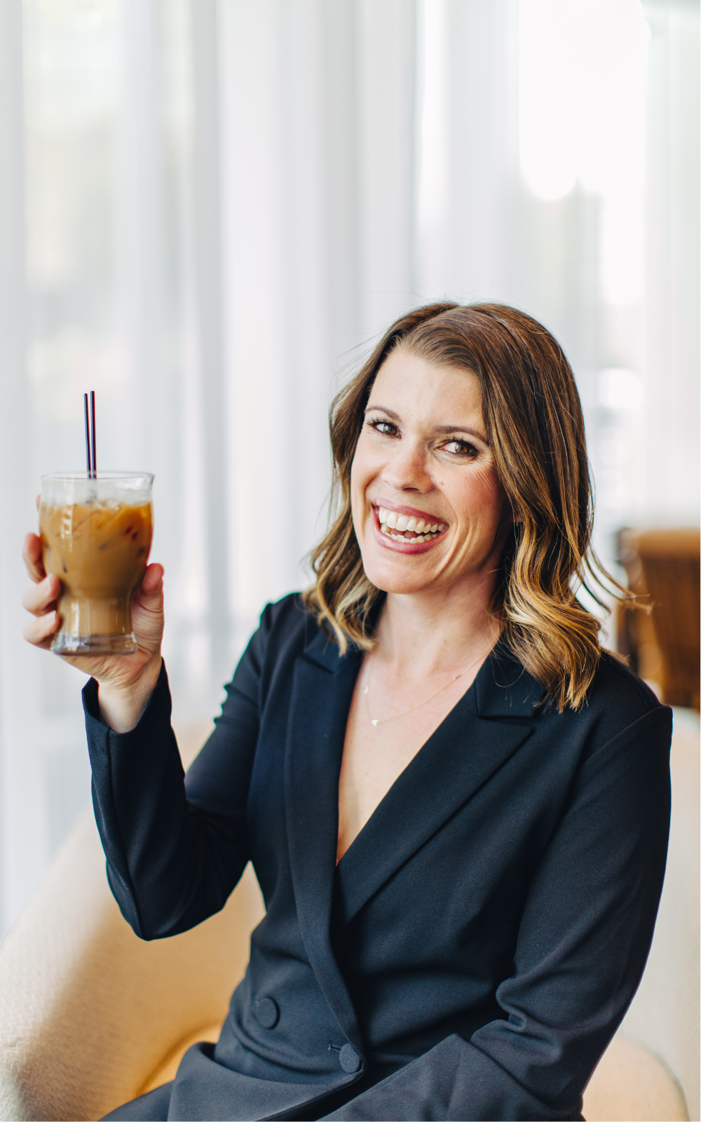 A woman with shoulder-length brown hair, smiling and holding a glass of iced coffee with black and red striped straws, sitting in a well-lit room with white curtains.