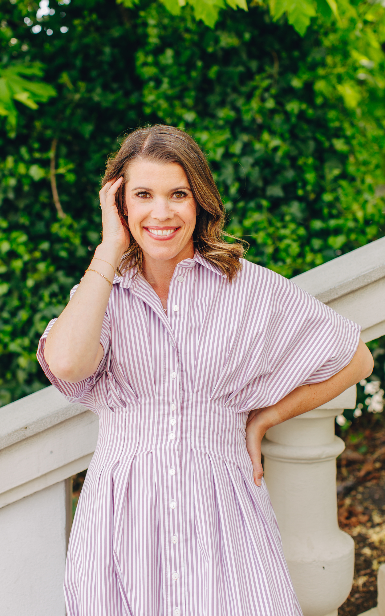 Smiling woman with brown hair wearing a pink and white striped dress, standing outdoors near a white stone railing with green foliage in the background.
