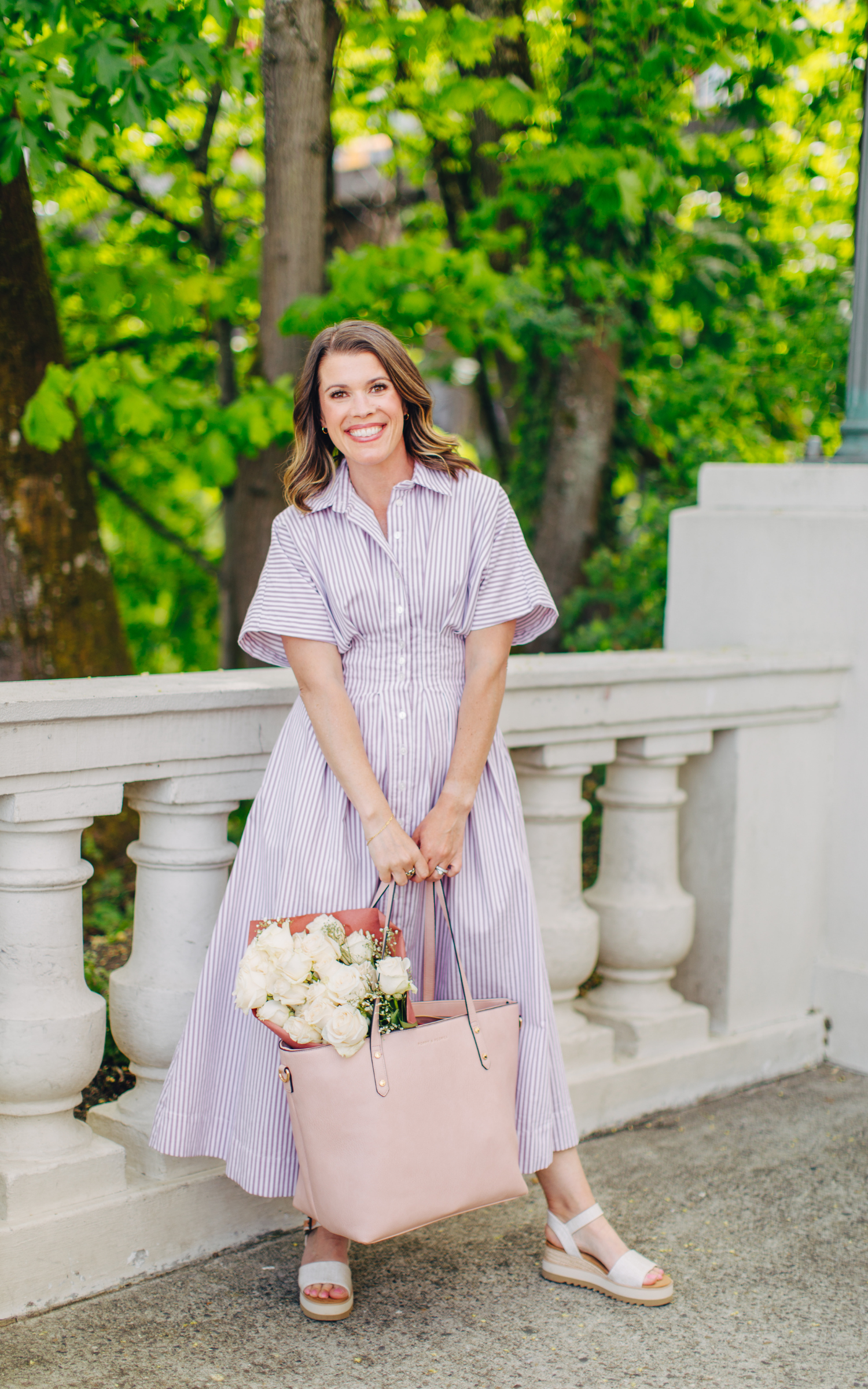 A woman in a purple and white striped dress smiling and holding a bouquet of white roses and a pink bag, standing outdoors beside a white railing with green trees in the background.