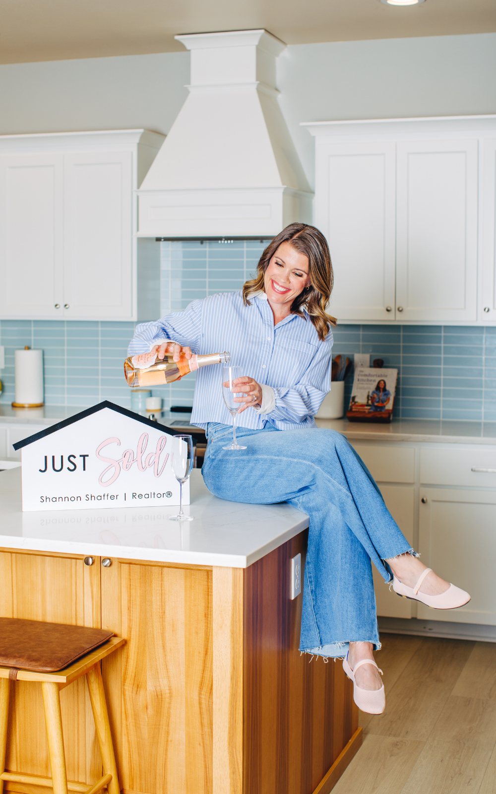A woman sitting on a kitchen island pouring champagne into a glass, celebrating a house sale, with a "Just Sold" sign in the foreground.