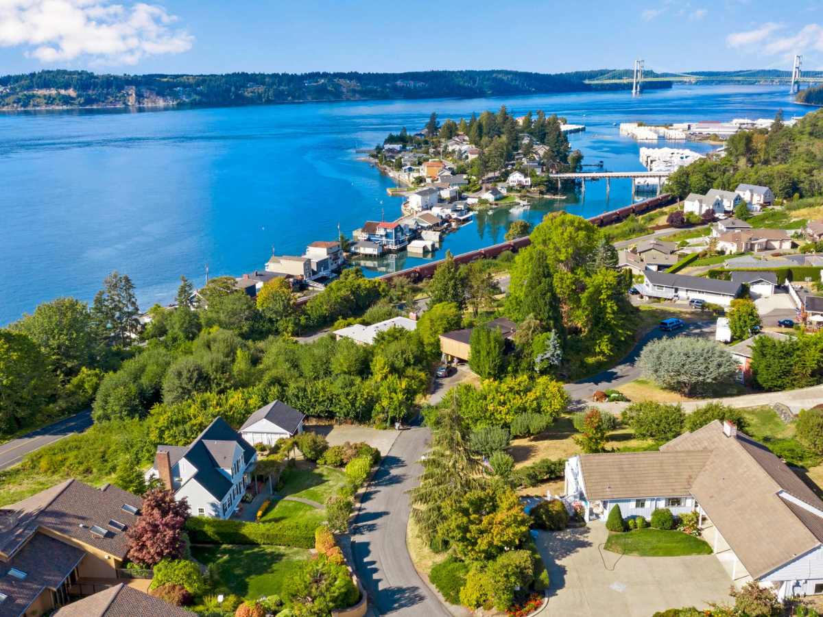 Aerial view of a coastal residential neighborhood with houses, trees, and a marina with boats, a train on tracks, and a large bridge over a body of water in the background.