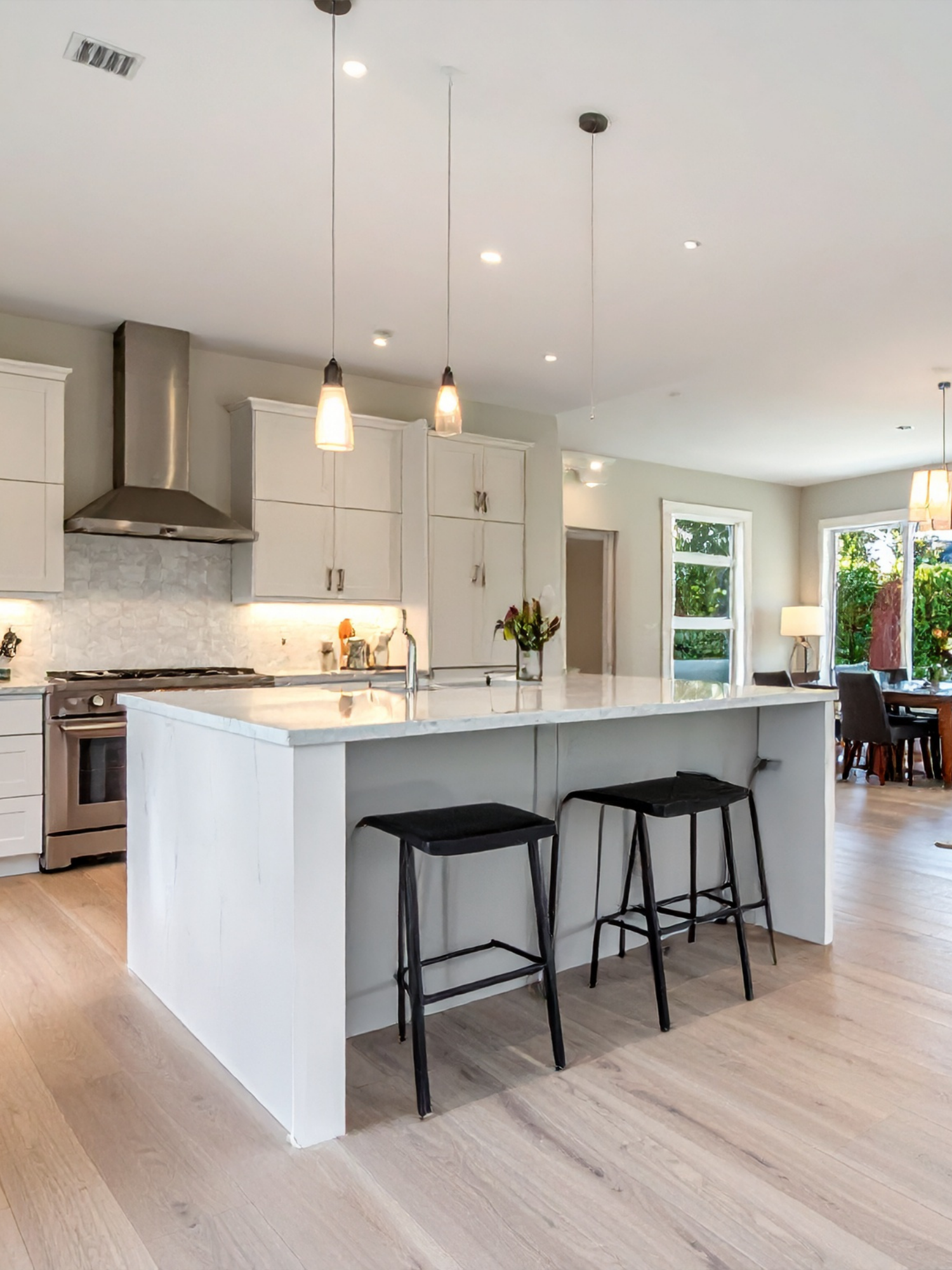 Modern kitchen with white cabinets, a marble kitchen island with black chairs, and pendant lighting, adjacent to a dining area with large windows and a table.