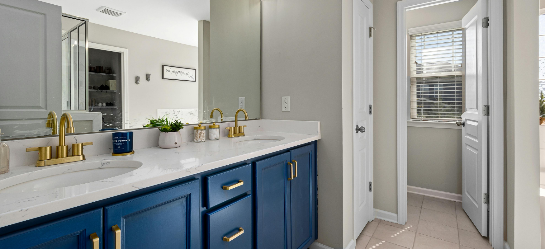 A modern bathroom with a double vanity featuring blue cabinets, gold handles, and a white marble countertop with two sinks and gold faucets. There are small plants and candles on the counter. A large mirror extends across the wall, reflecting a walk-in closet with shoes. To the right, there are two doors leading to a small laundry area with a window and blinds, illuminated by natural light.