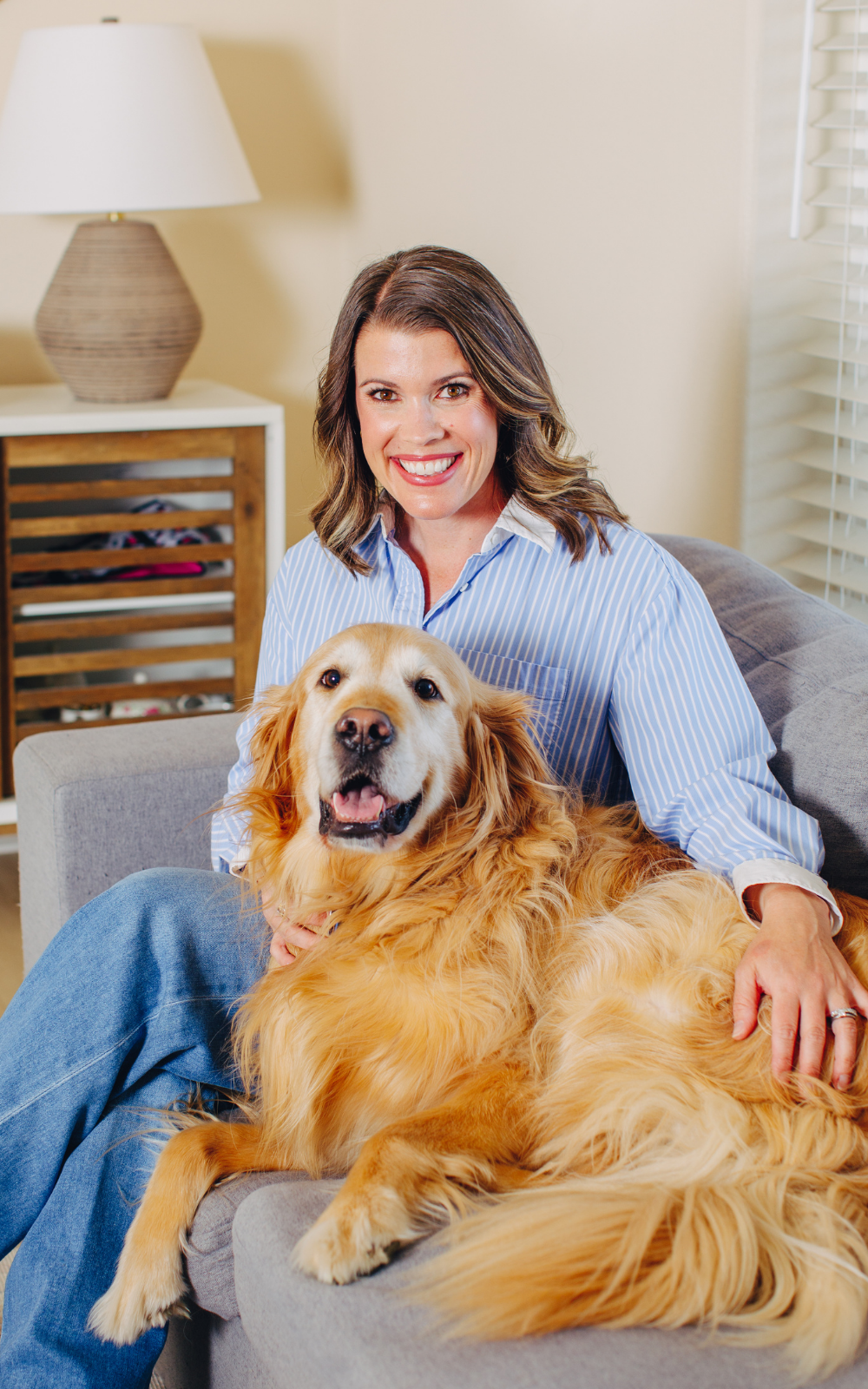 A woman with shoulder-length brown hair smiling at the camera, sitting on a gray couch with her golden retriever dog, inside a well-lit living room with beige walls, a white lamp, and a window with blinds.