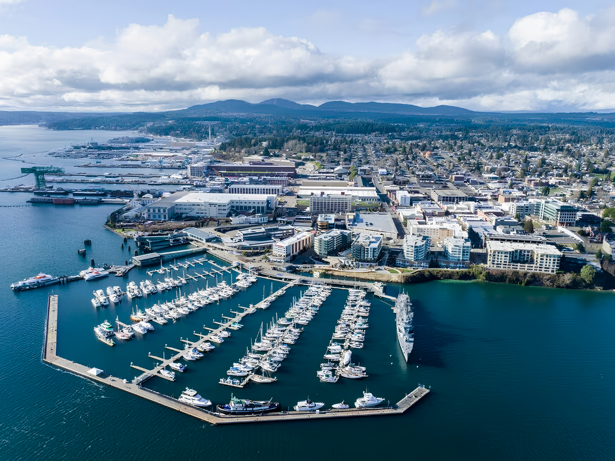 Aerial view of a city with a marina filled with boats and yachts, buildings, and a harbor, with mountains and cloudy sky in the background.