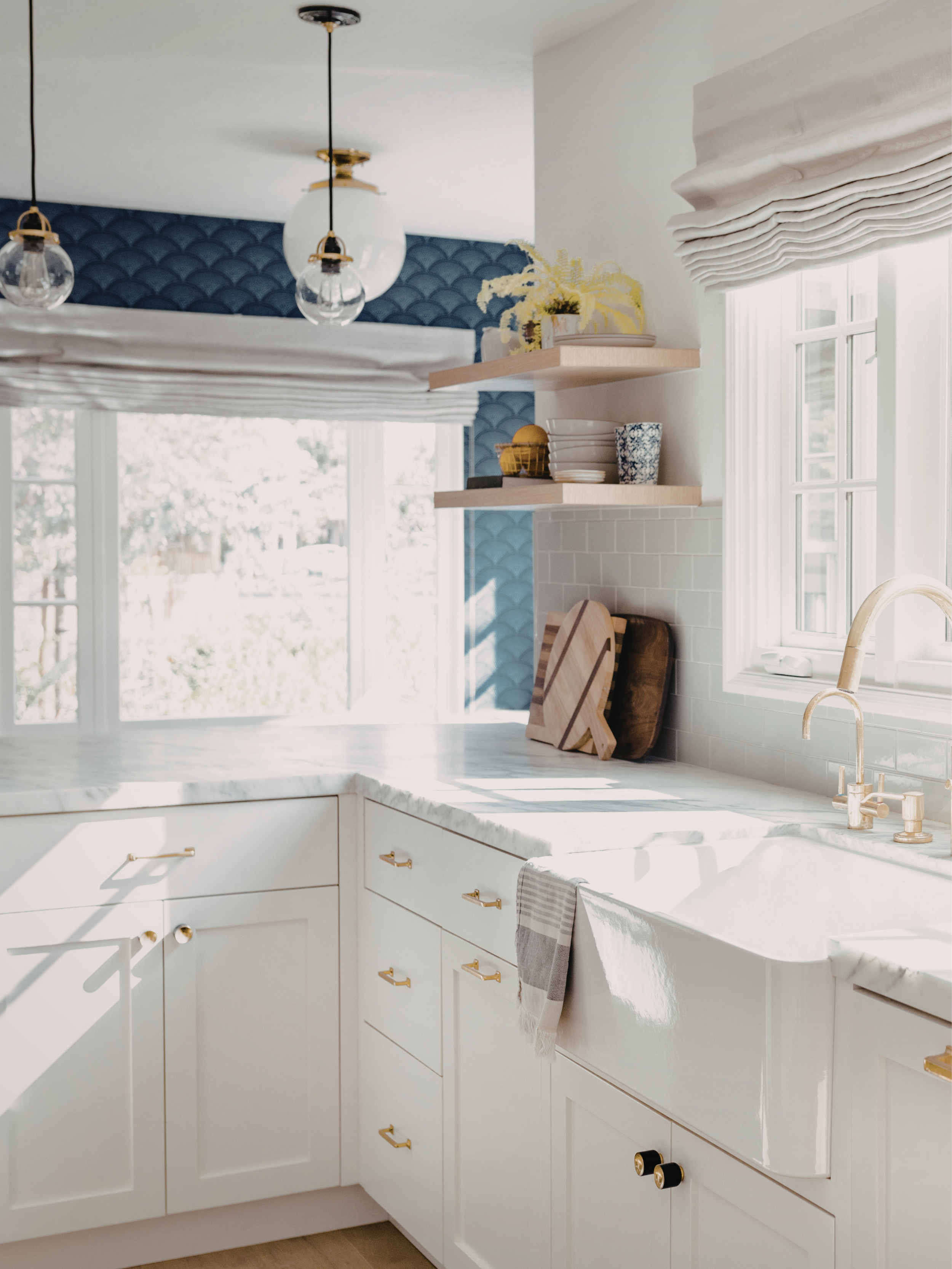 A bright kitchen with white cabinets, a marble countertop, a window with Roman shades, open shelves with dishes and decor, a gold faucet, and natural light.
