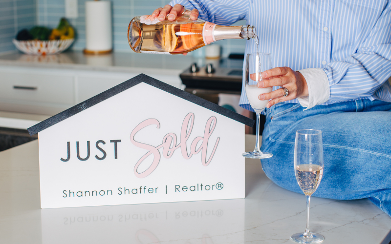 Person pouring champagne into a glass in front of a house-shaped sign that says 'JUST sold' by Shannon Shaffer, Realtor.