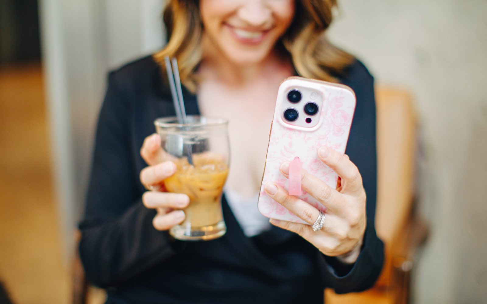 Woman smiling and looking at her phone while holding a glass of iced coffee with two straws.