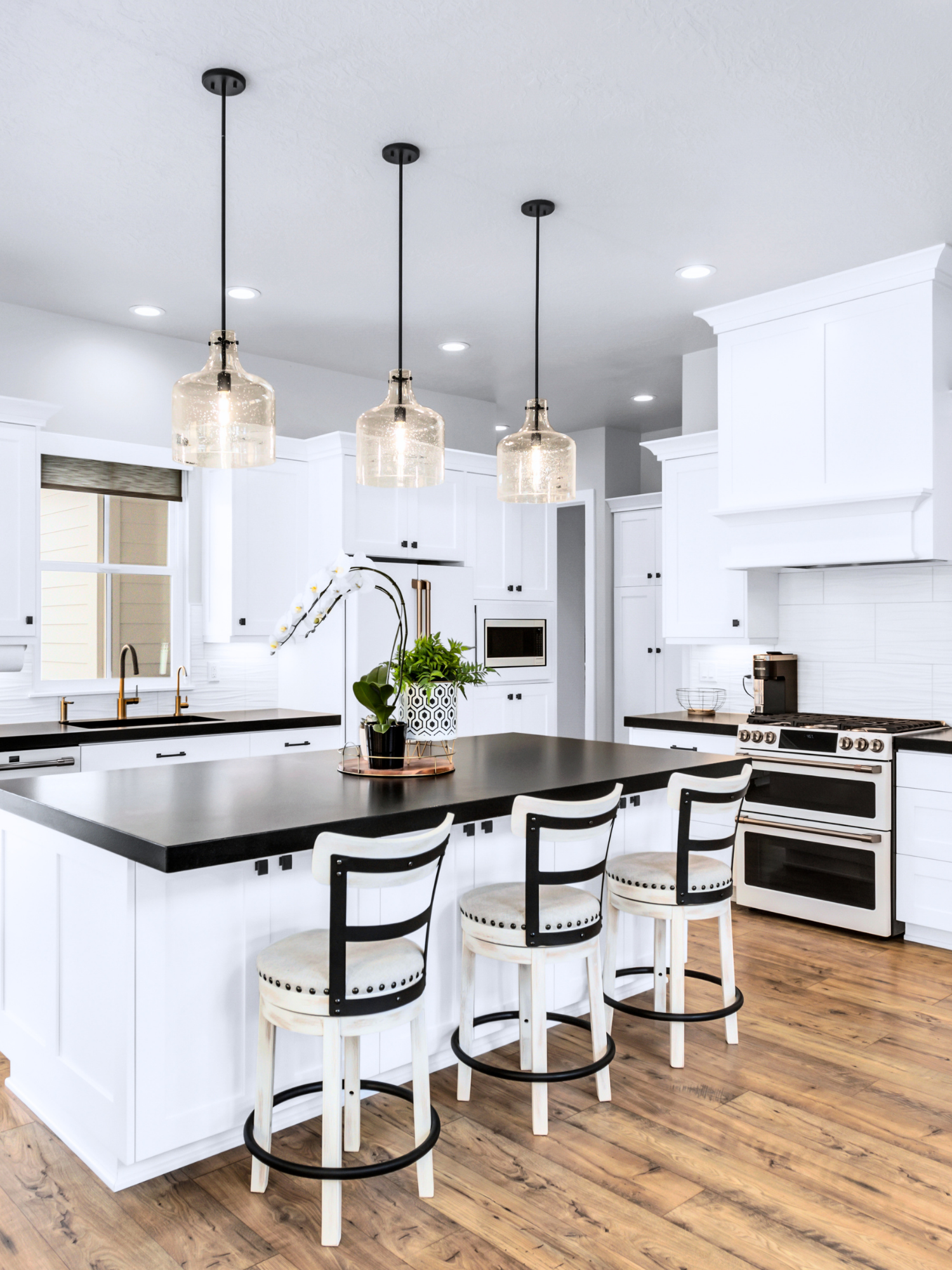 Modern kitchen with white cabinets, black countertops, and a wooden floor. Four white chairs with black accents are arranged around an island with a black surface. Three pendant lights hang from the ceiling above the island. There is a potted plant and orchids on the island, a window with a partially closed blind, and built-in appliances including a microwave and oven.
