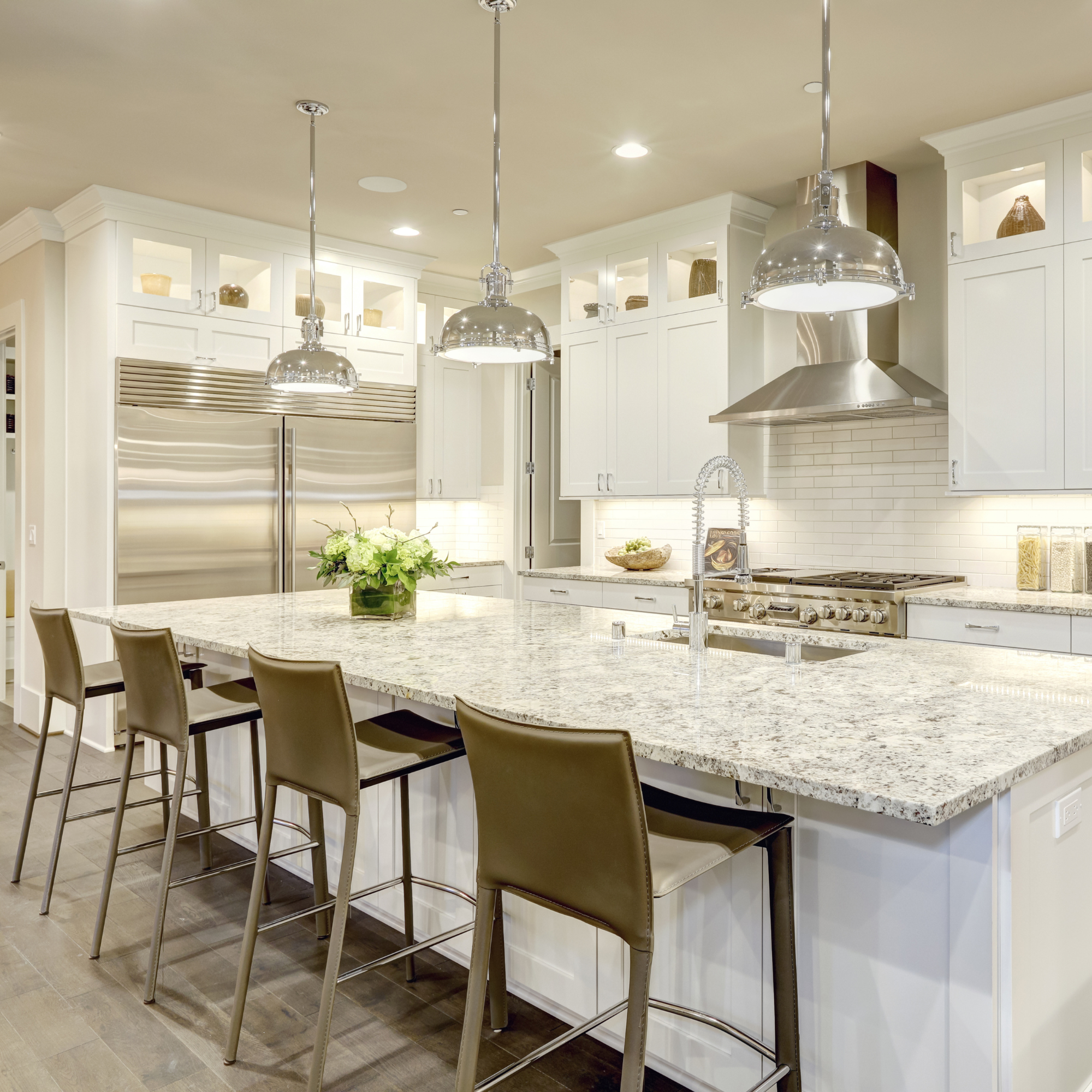 Modern kitchen with white cabinets, a large island with a granite countertop, three metal barstools, stainless steel appliances, pendant lights, and a flower arrangement.